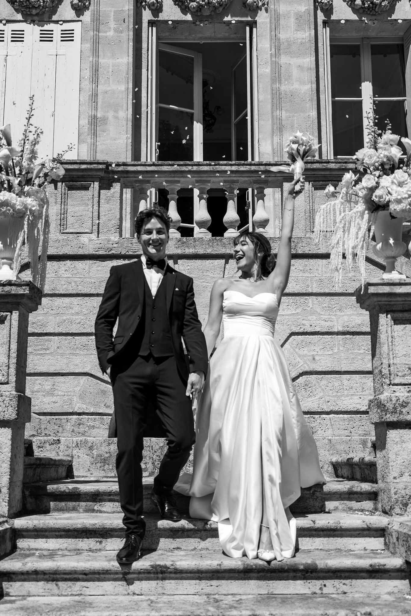 Black and white photo of bride and groom descending chateau stone steps with confetti in the air after ceremony