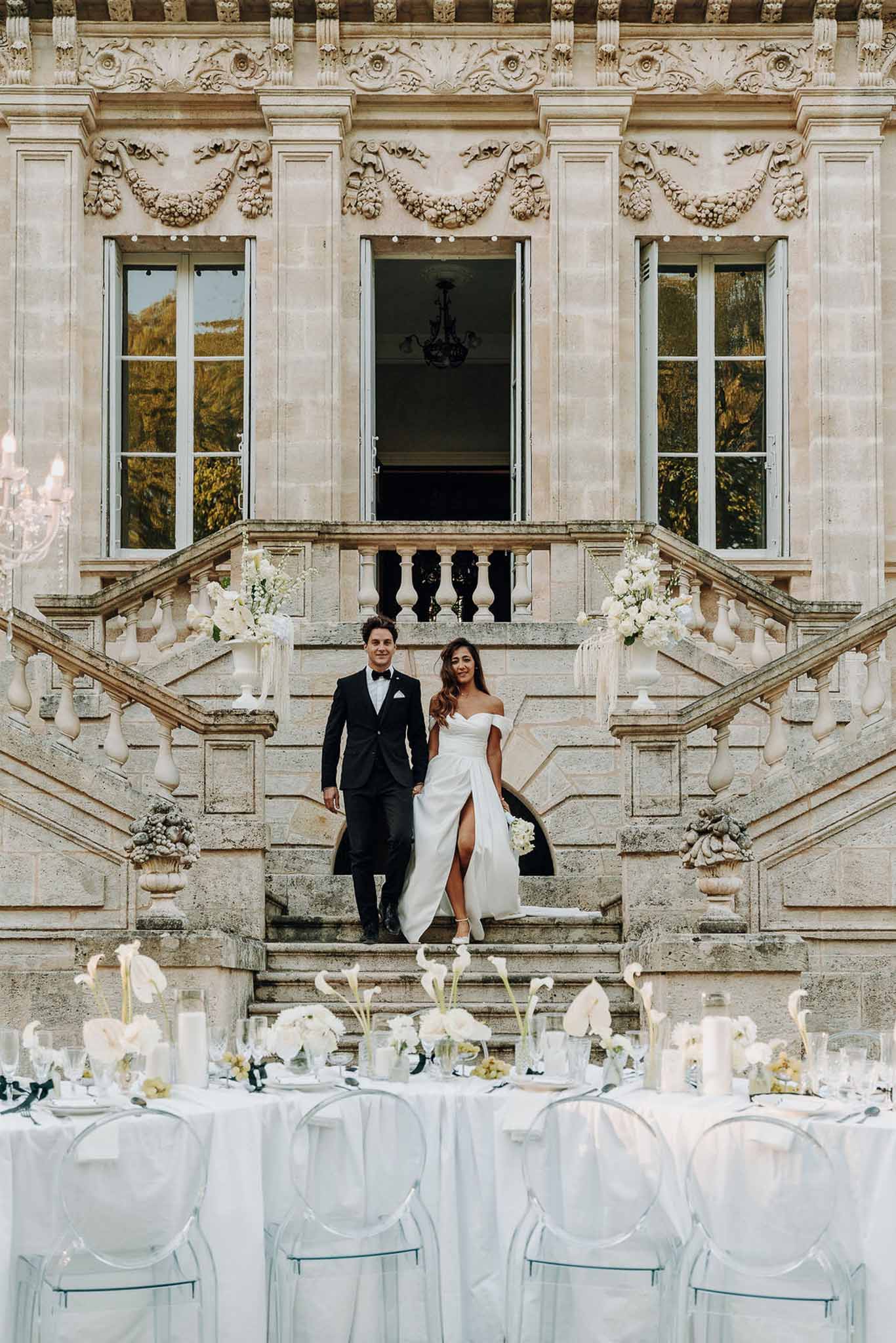 Couple descending grand chateau staircase toward all-white reception table with ghost chairs calla lilies and candles