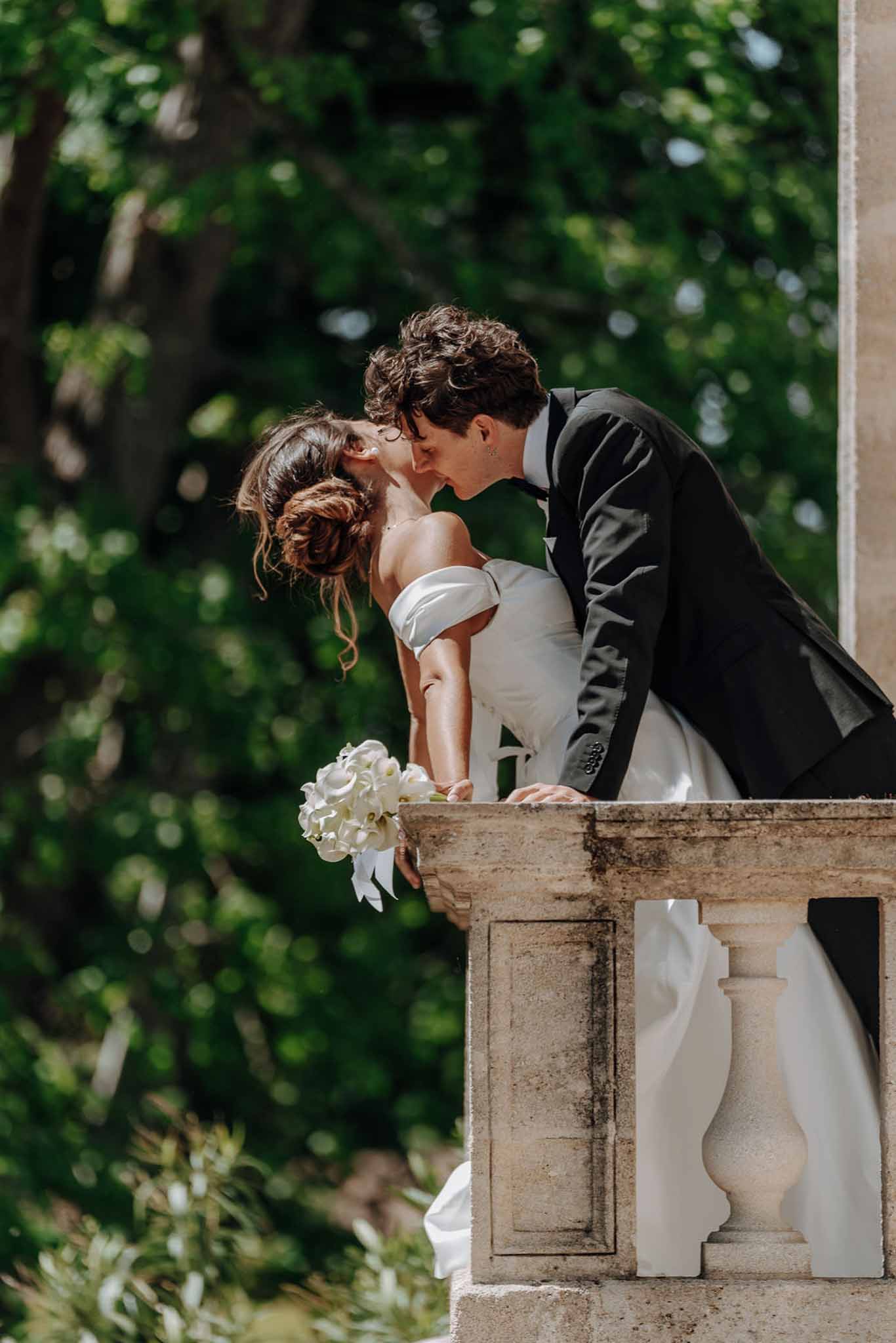 Couple kissing on stone balustrade balcony from below, bride holding white calla lily bouquet with green foliage behind