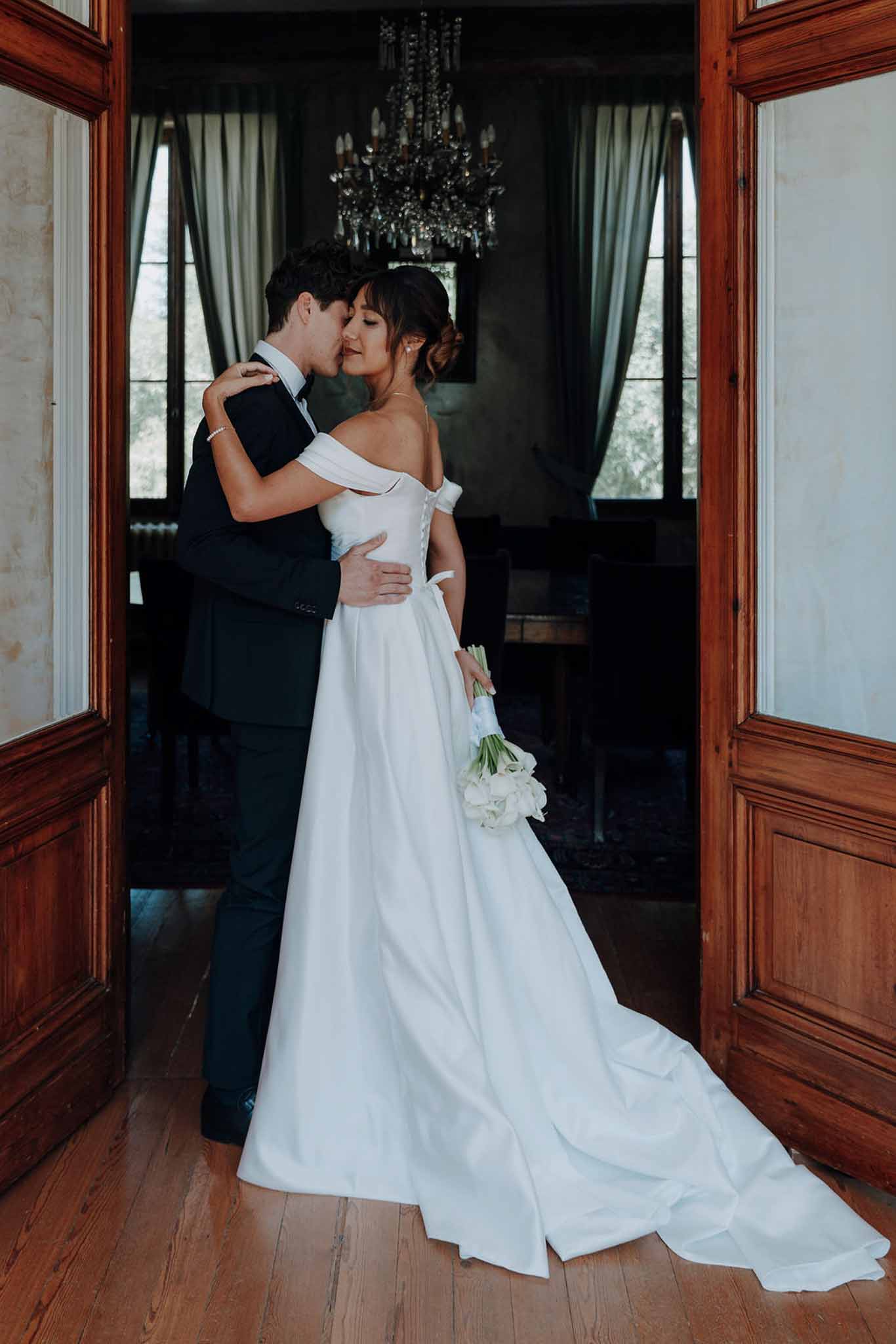 Groom in navy suit kissing bride in off-the-shoulder gown with calla lily bouquet in chateau doorway