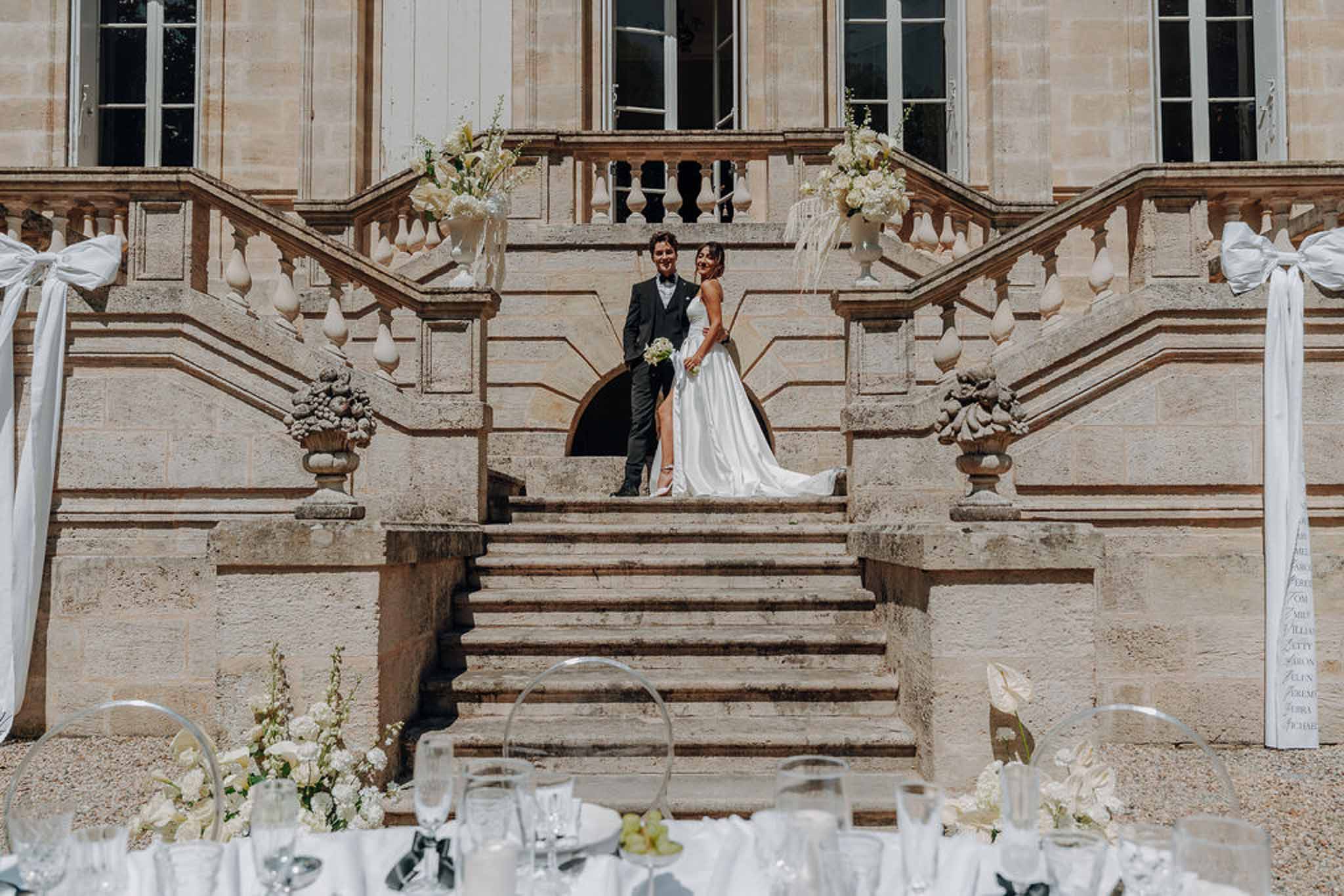 Couple on grand chateau staircase with white floral urns, ghost chair reception tables in foreground