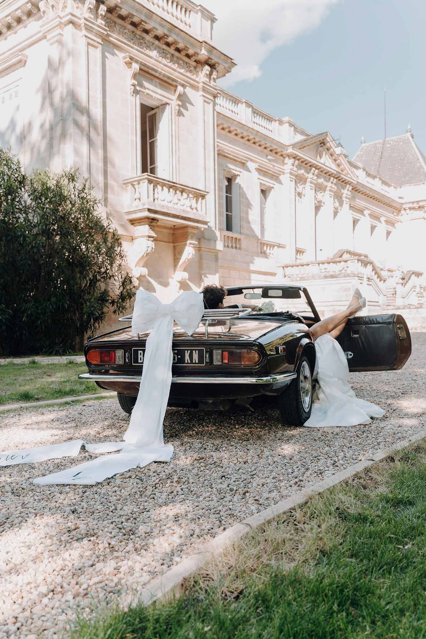 Bride with legs kicked up in black vintage convertible and groom driving before carved chateau facade