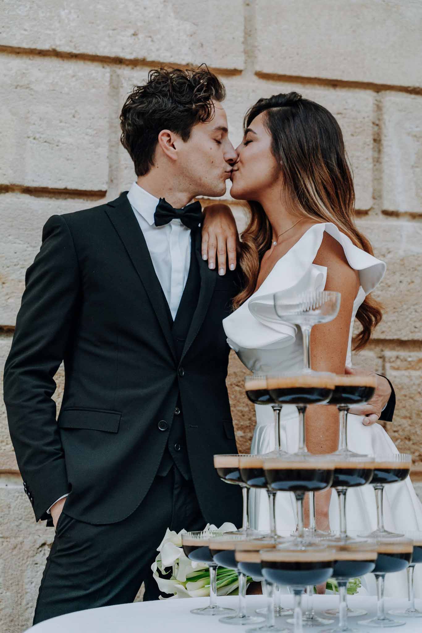 Couple kissing behind coupe glass tower filled with red wine against stone chateau wall