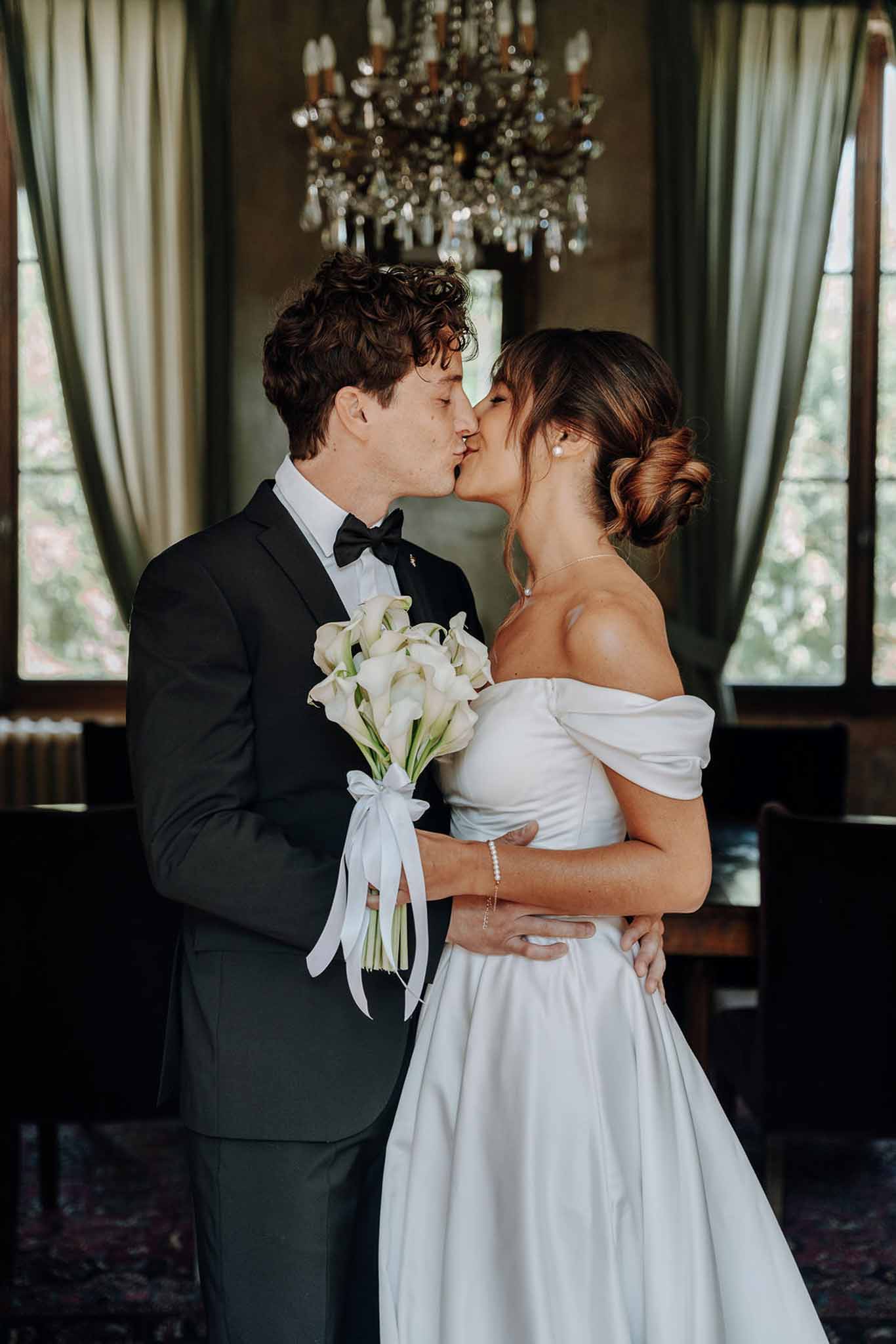 Bride and groom kissing beneath crystal chandelier in chateau reception room with calla lily bouquet