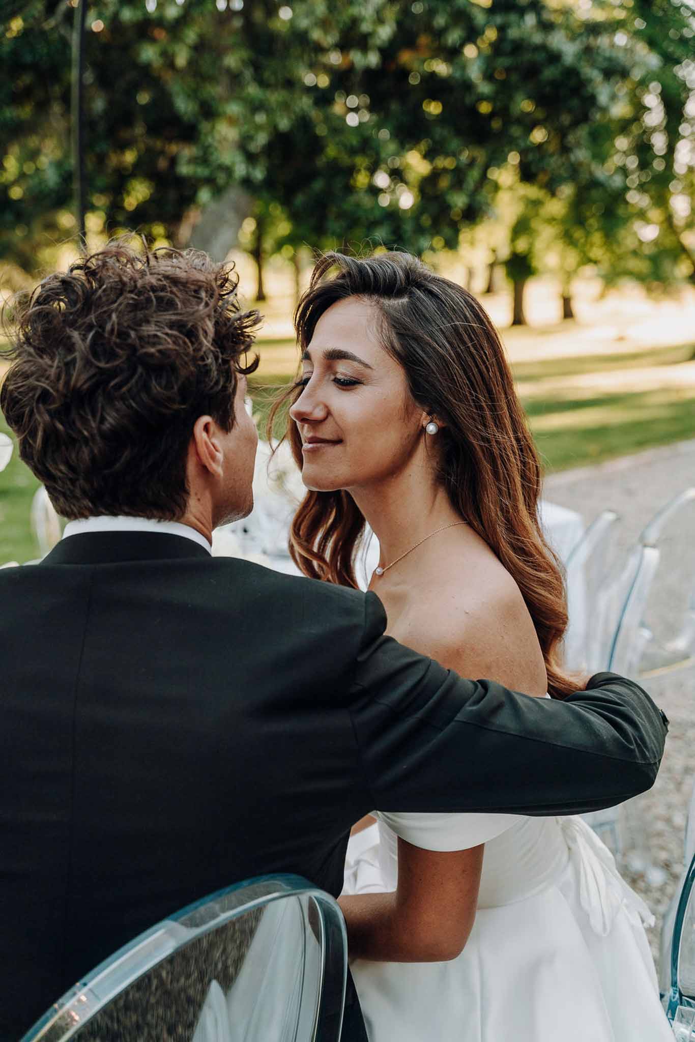 Close-up over groom's shoulder of smiling bride with pearl earrings in off-shoulder gown at golden hour