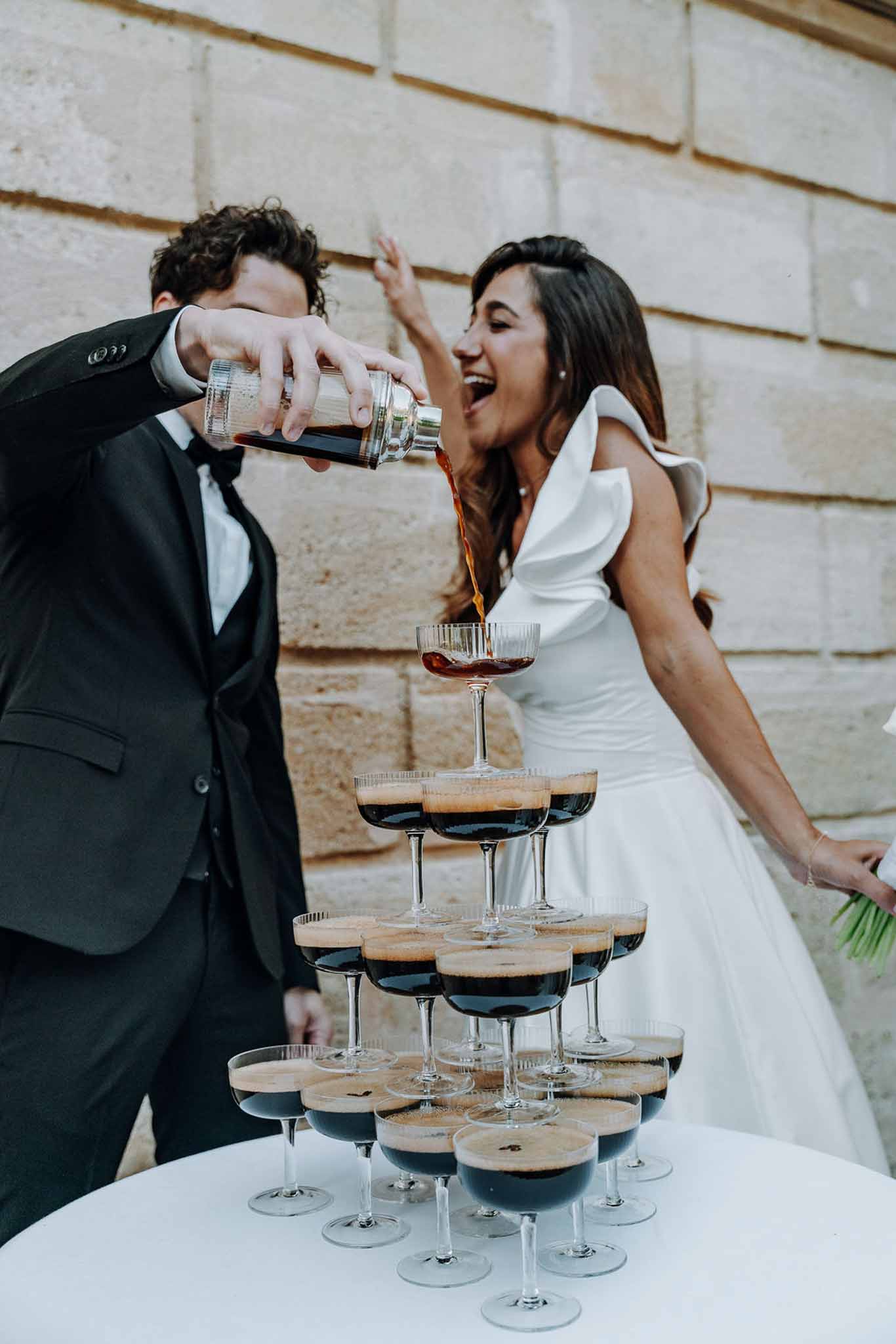 Bride and groom pour espresso martinis into a coupe glass tower against a stone wall outdoors