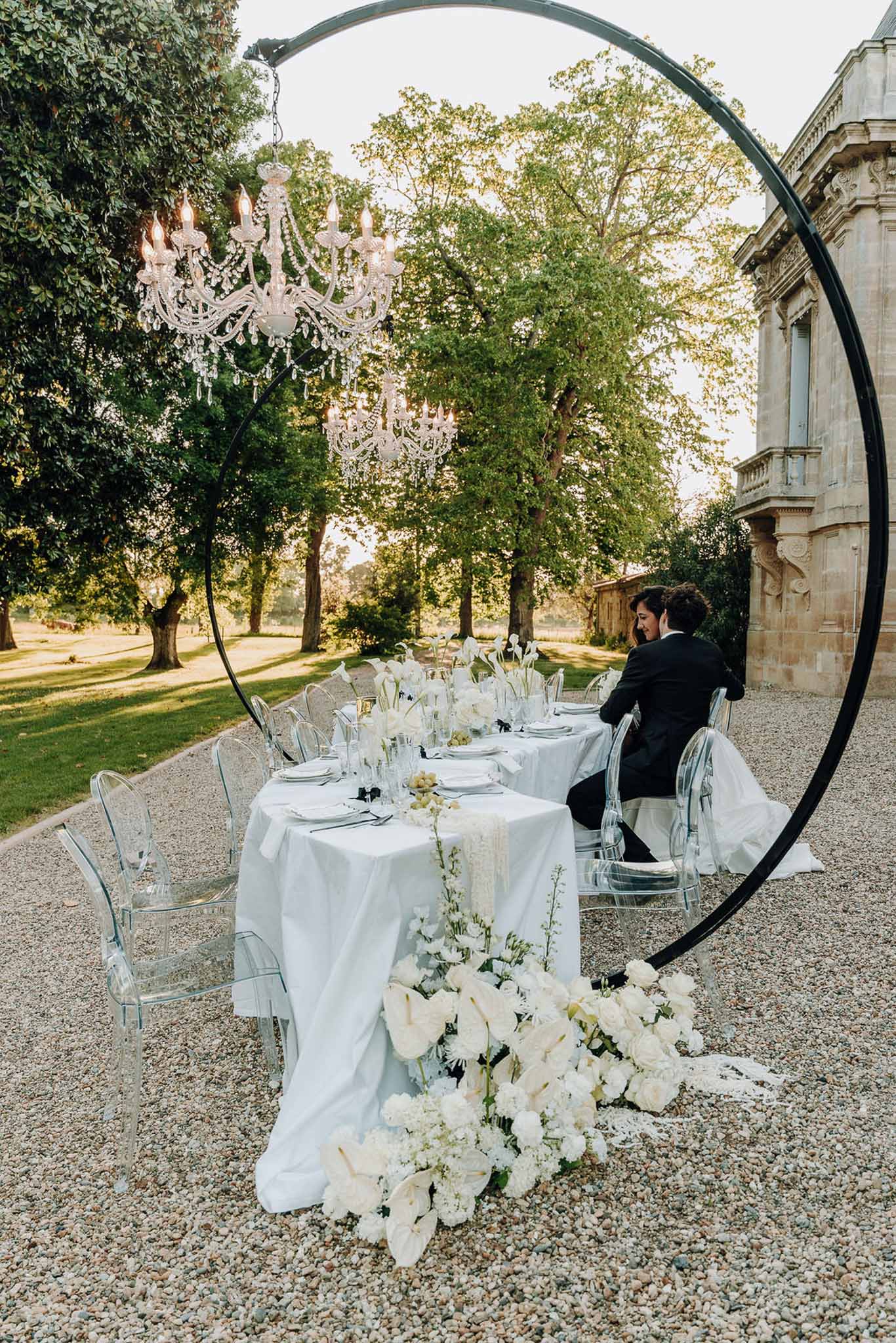 Monochrome reception table with ghost chairs, white calla lily runner, and black circular arch with crystal chandeliers