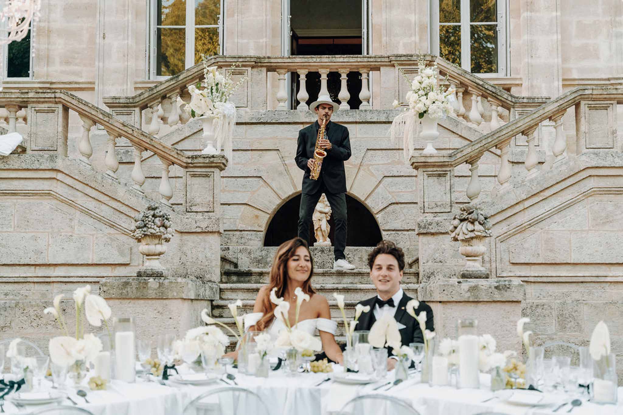 Saxophonist on staircase behind couple at sweetheart table with white calla lily and orchid arrangements