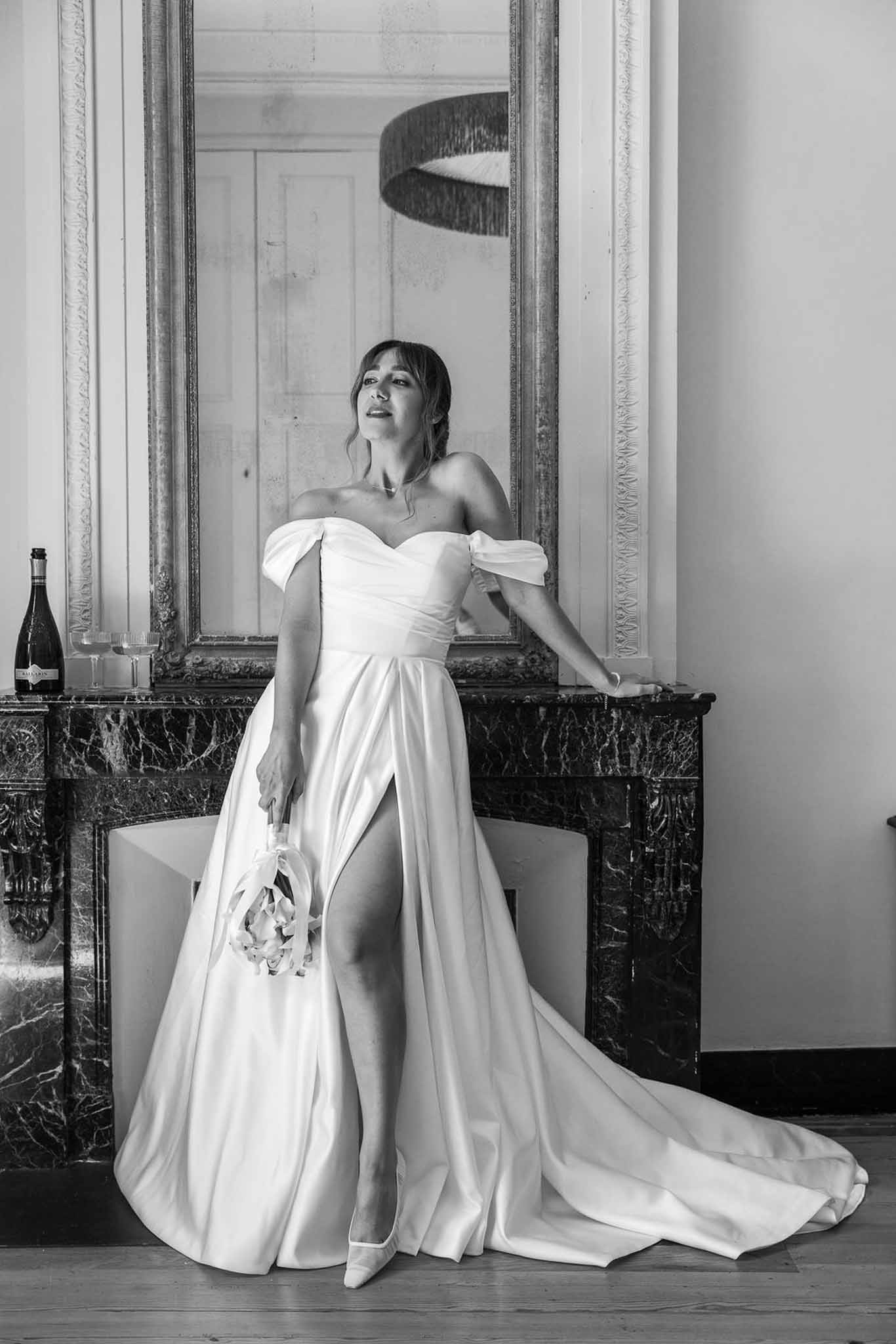 Black and white full-length portrait of bride in off-shoulder satin ball gown with slit beside marble fireplace