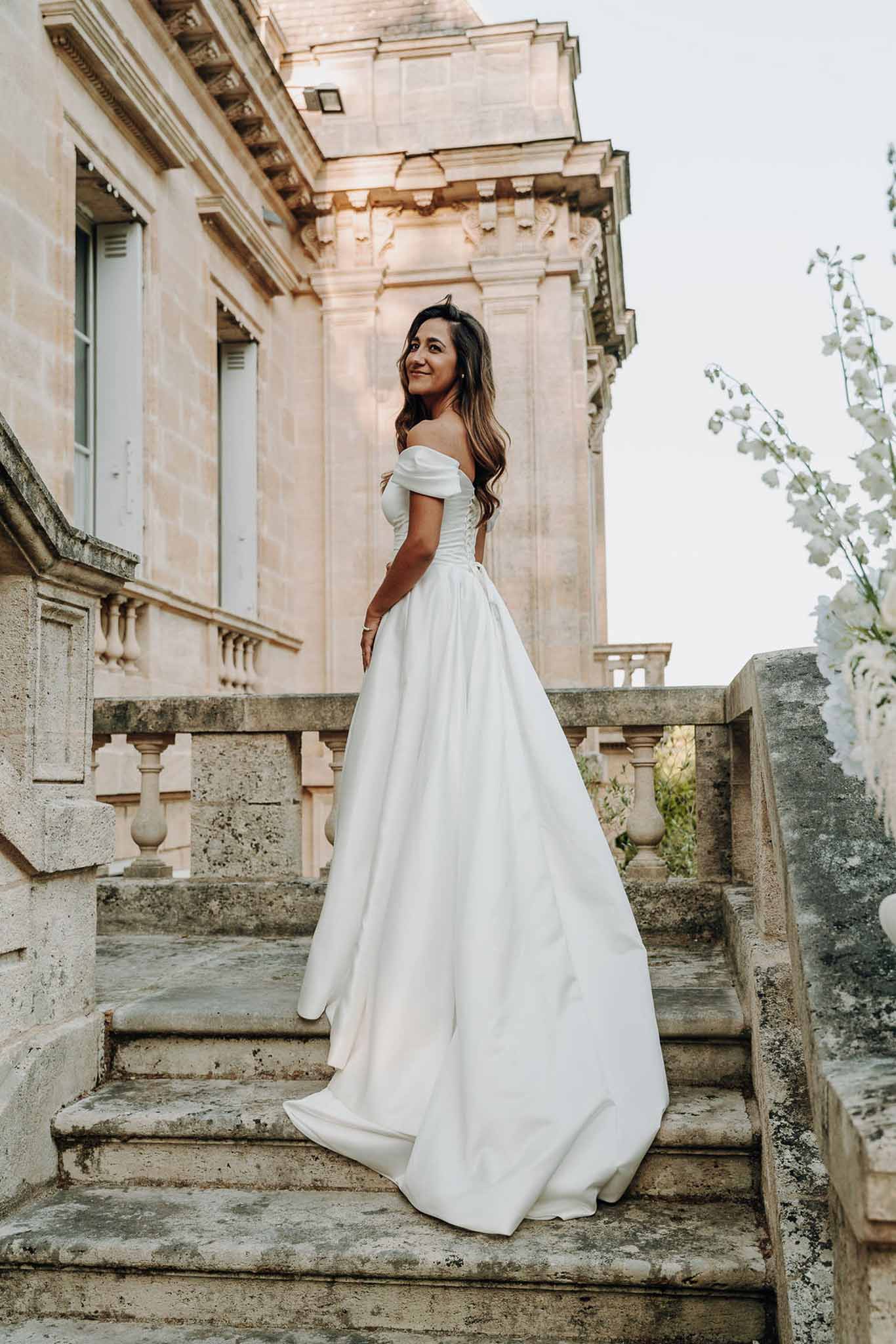 Bride glancing over shoulder on chateau staircase with off-shoulder ballgown train on steps
