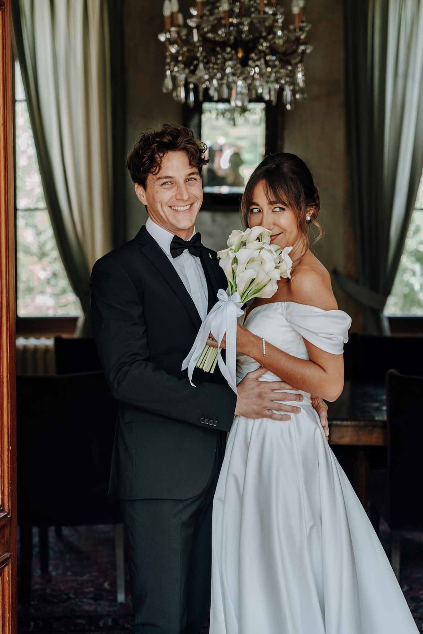 Couple in chateau interior bride in off-shoulder satin ballgown with calla lily bouquet groom in tuxedo chandelier behind