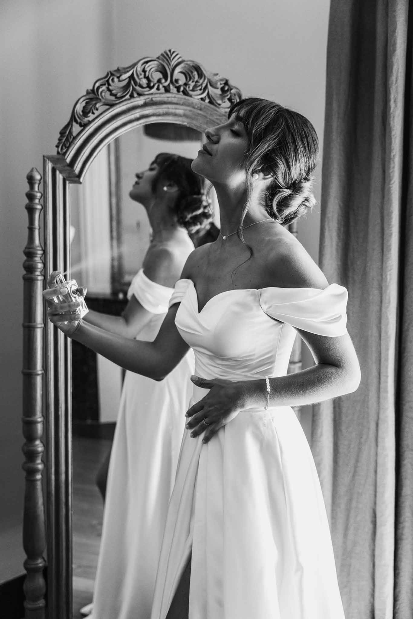 Black and white bride applying perfume before ornate floor mirror wearing off-shoulder A-line gown and chignon