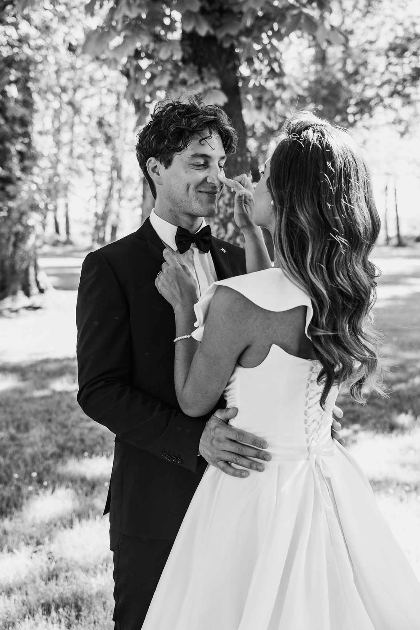 Bride playfully touching groom's nose in backlit wooded park setting in black and white