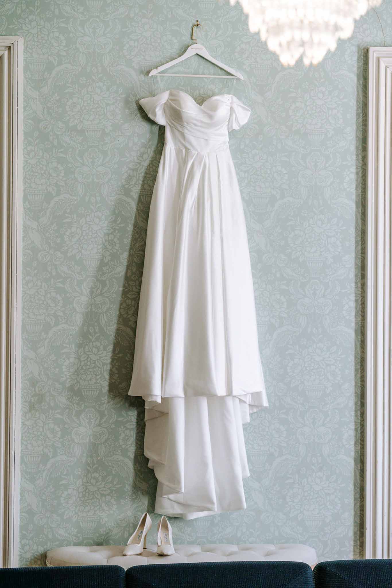 White off-the-shoulder bridal gown hanging on sage floral wallpaper with white heels on tufted bench below
