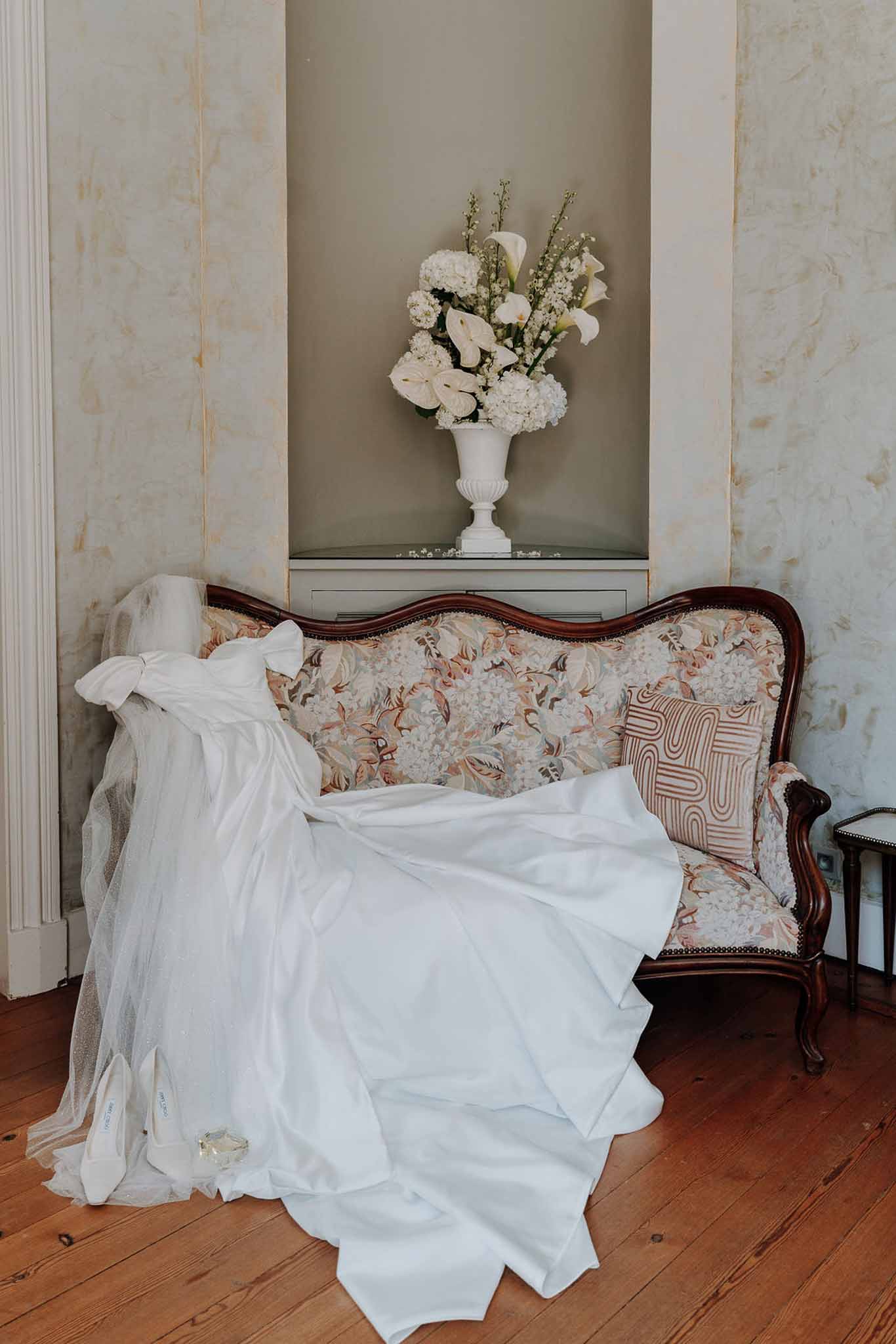 Wedding gown with bow detail draped on carved French sofa beside white heels, veil, and hydrangea urn