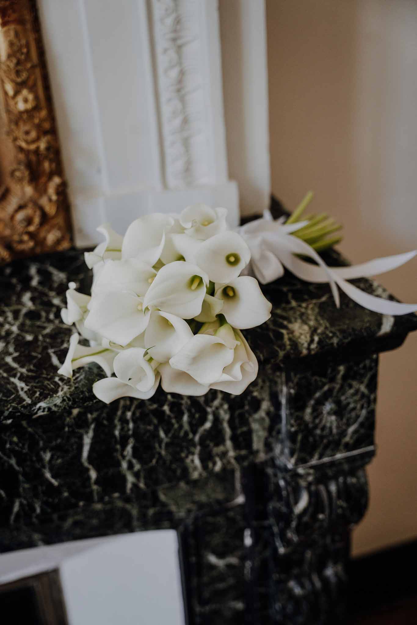 White calla lily bridal bouquet with satin ribbon on green marble fireplace mantel in formal chateau interior