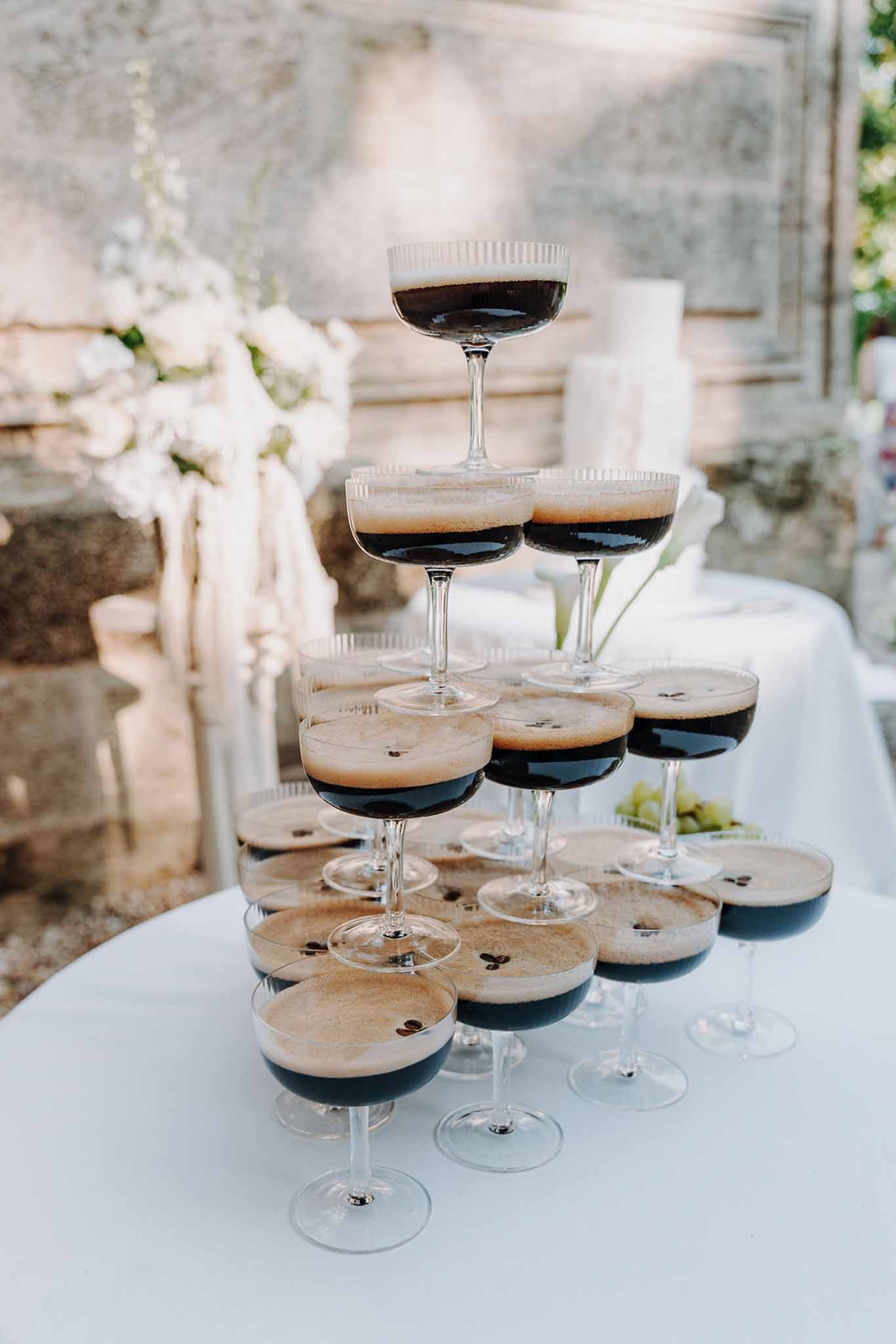 Espresso martini coupe pyramid with coffee bean garnish on white linen beside white floral arrangement