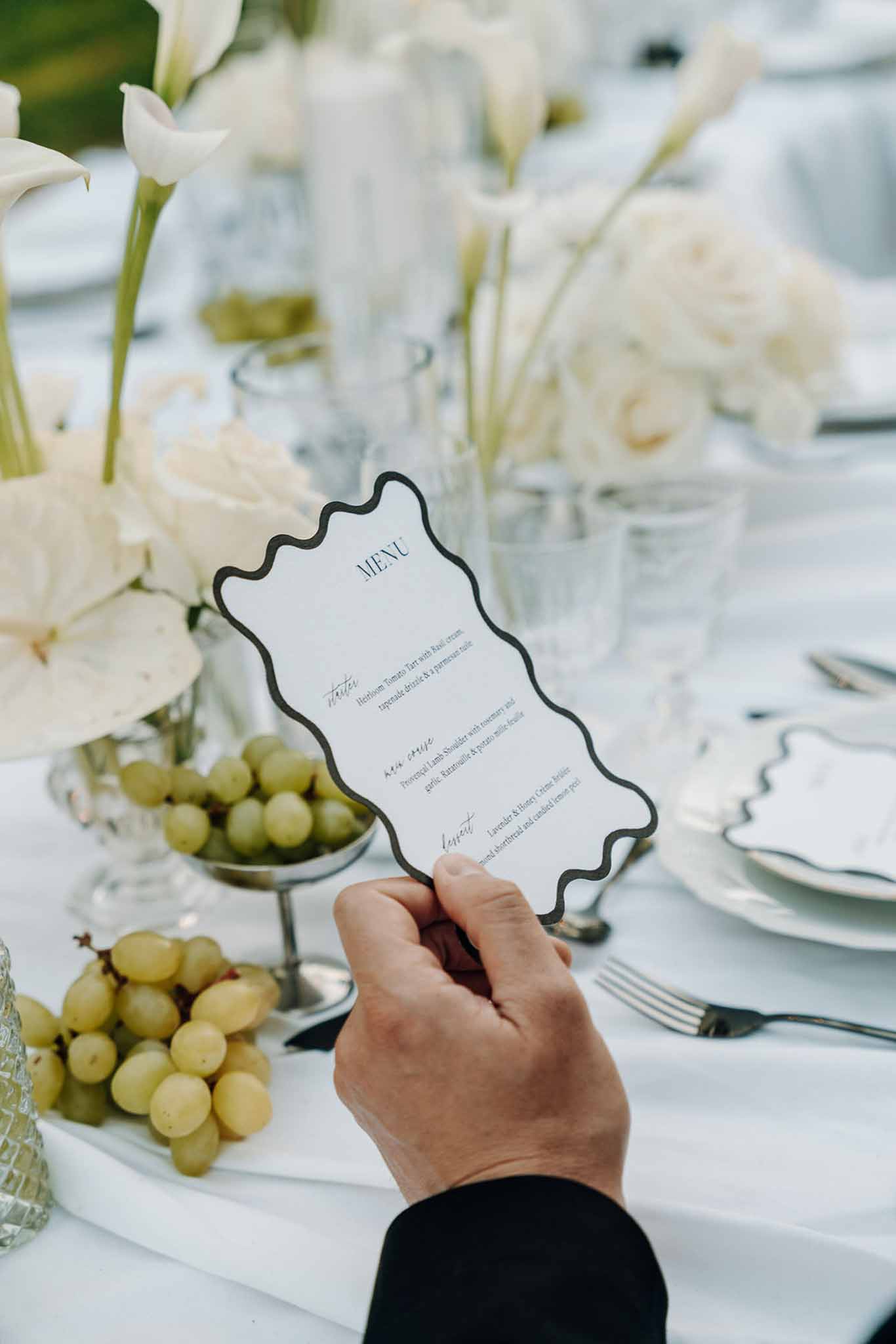 Hand holding wavy-bordered menu card at monochrome table with calla lilies, peonies, and green grapes