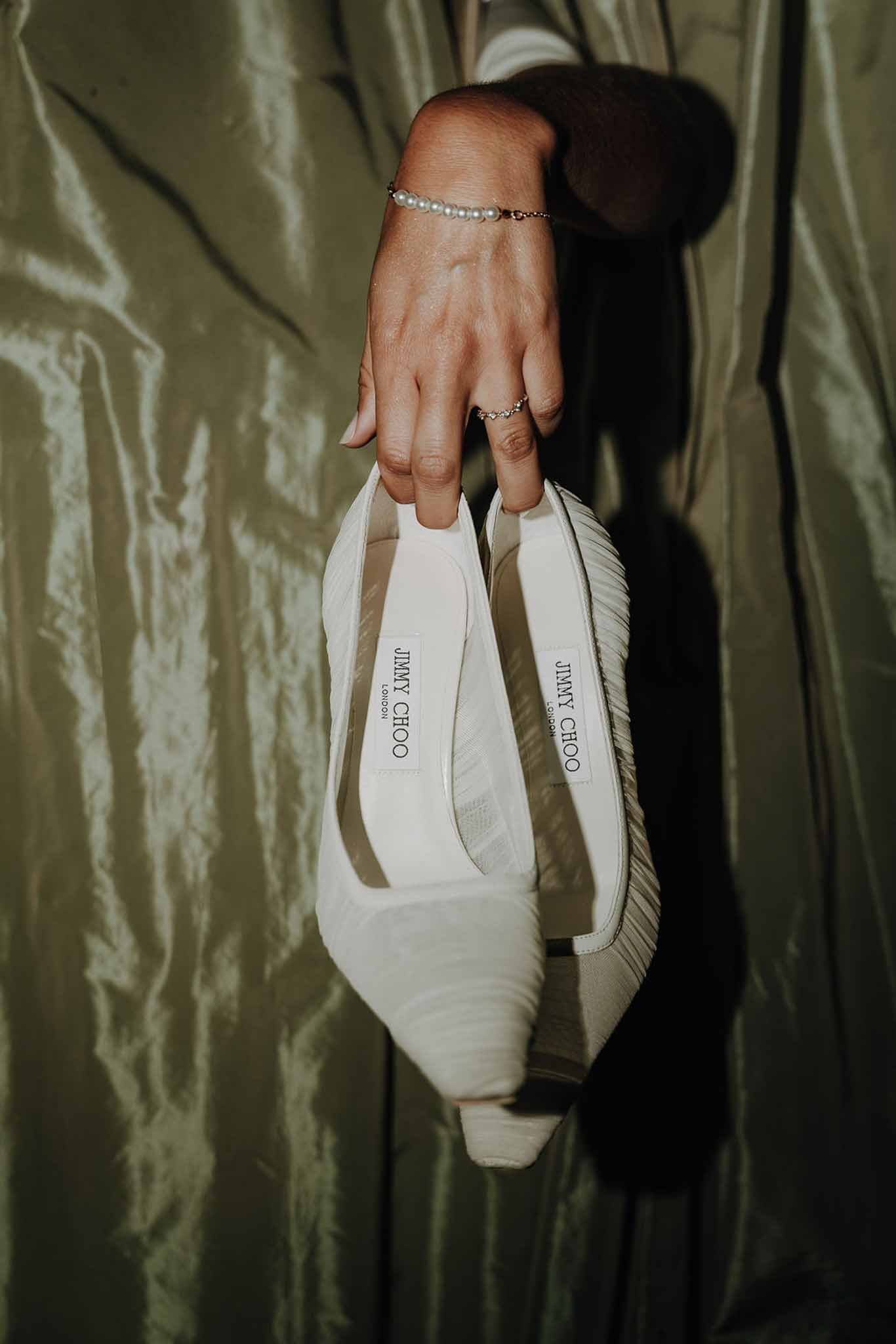 Bride holding ivory Jimmy Choo heels with pearl bracelet against olive-green satin fabric backdrop