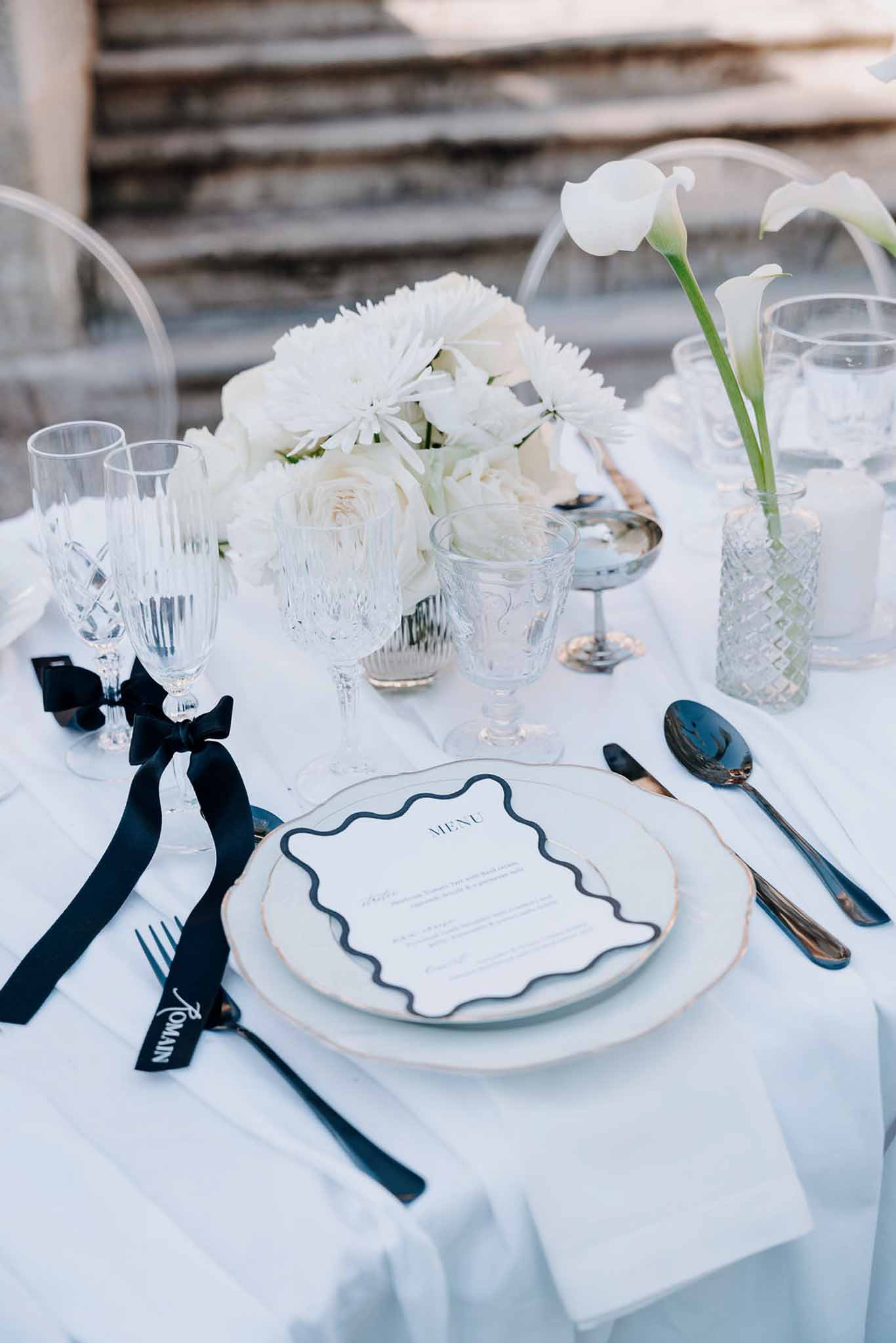 Monochrome table setting with black cutlery, wavy-border menu card, white roses, and ribbon-tied champagne flutes