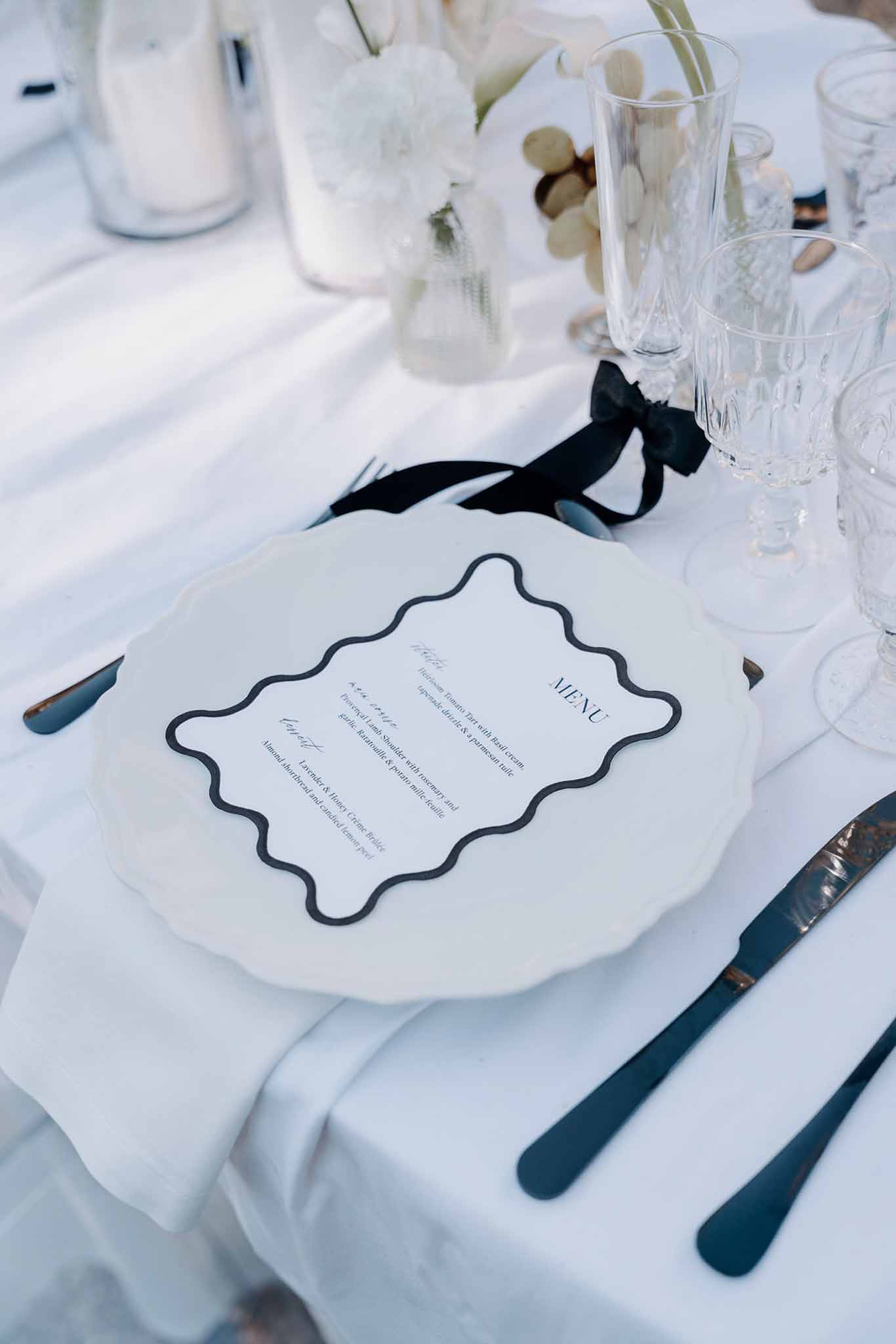 Monochrome place setting with wavy-bordered menu card, black ribbon, dark cutlery, and white florals