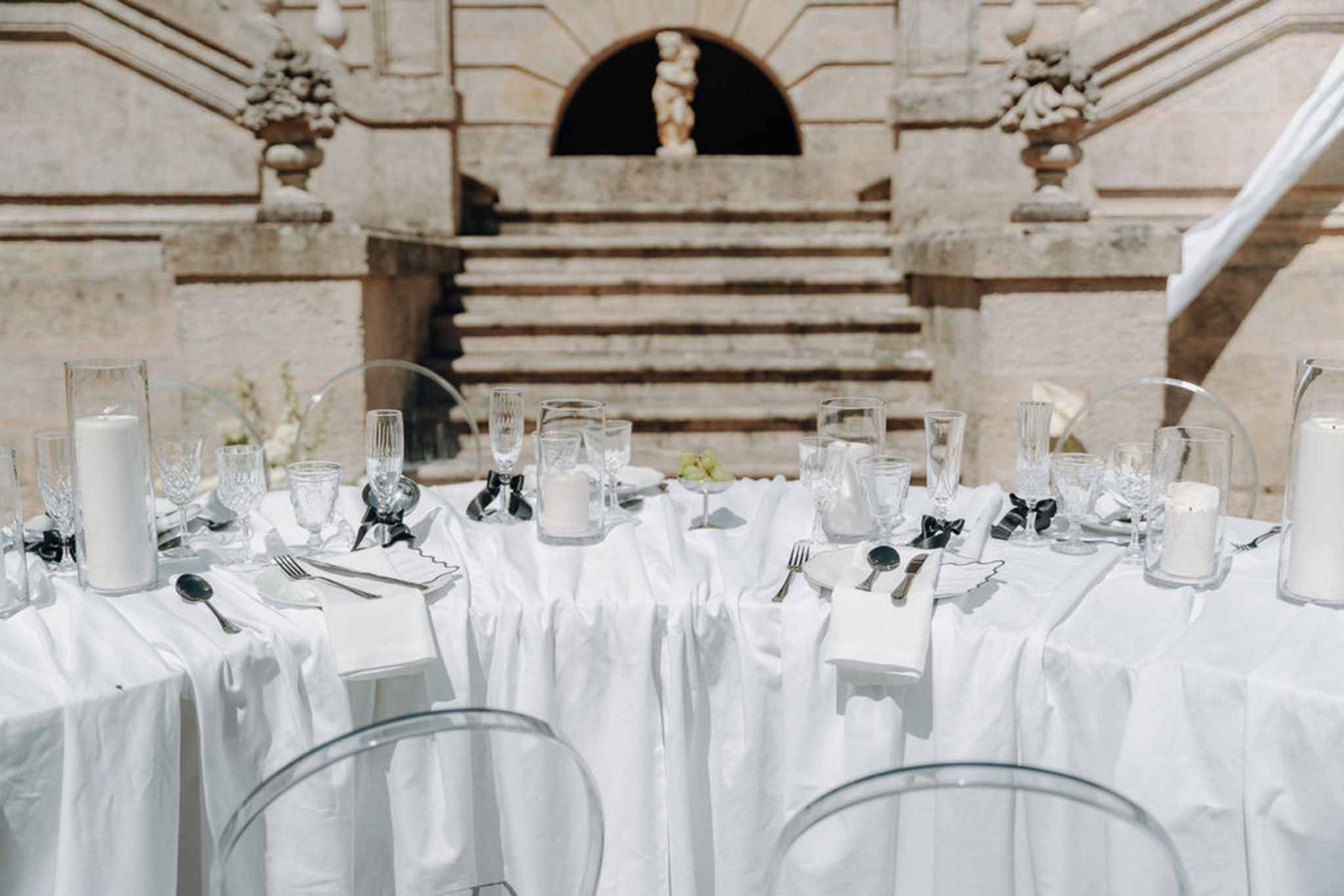 Round white table with ghost chairs, pillar candles, and black bow napkin rings before classical stone staircase niche