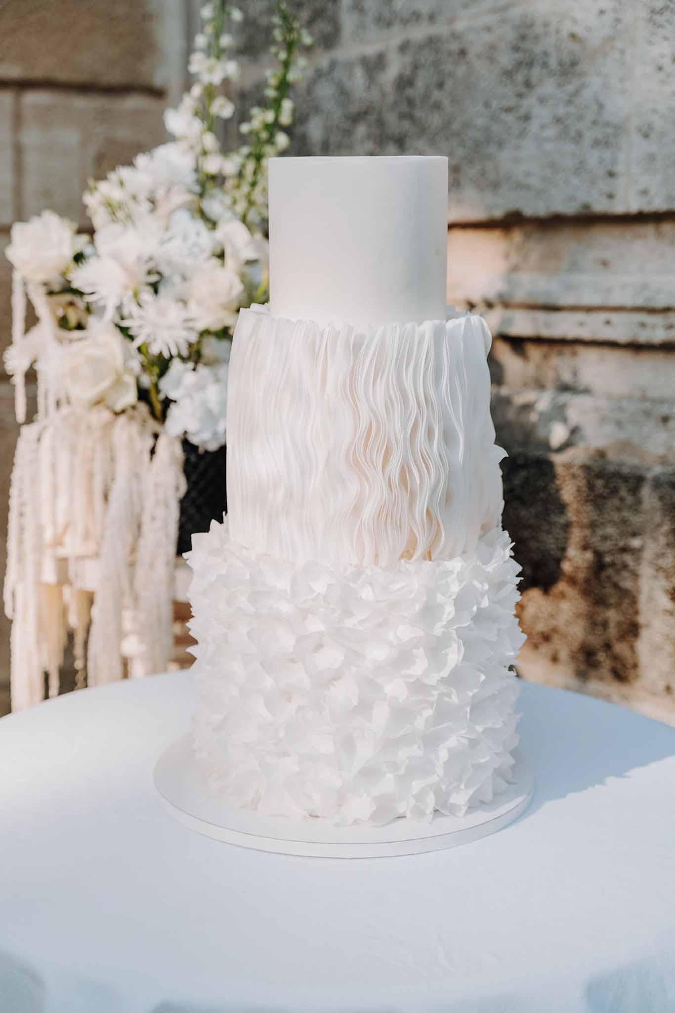 Three-tier white wedding cake with textured sugar ruffles and white floral arrangement against stone wall