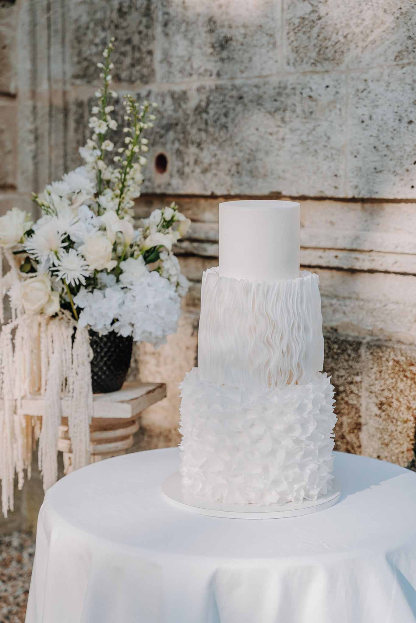 Four-tier all-white wedding cake with ruffled fondant petals on a linen-covered table beside white floral arrangement