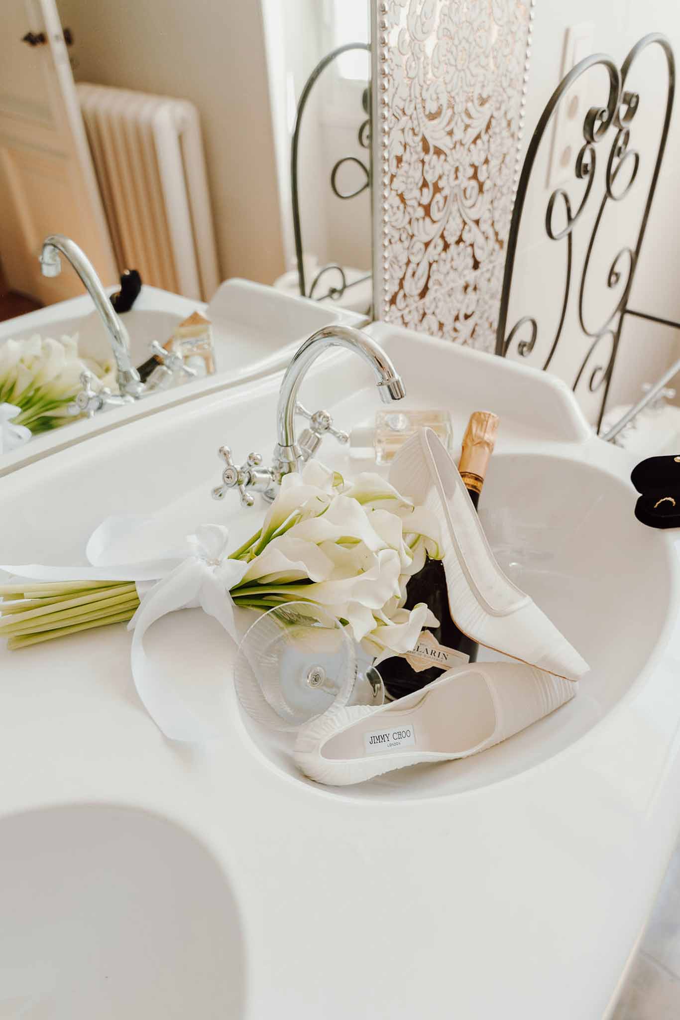 Jimmy Choo heels, calla lily bouquet, champagne, and ring box arranged in white ceramic double basin sink