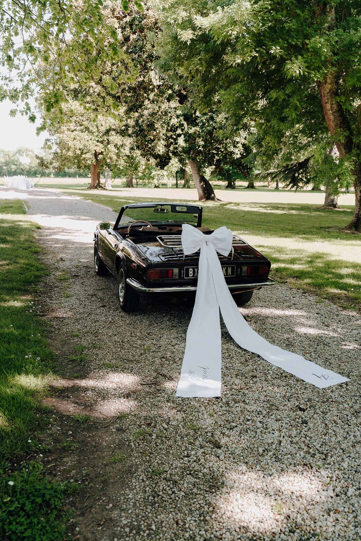 Dark vintage convertible car decorated with a large white bow and long trailing ribbon on a chateau gravel driveway