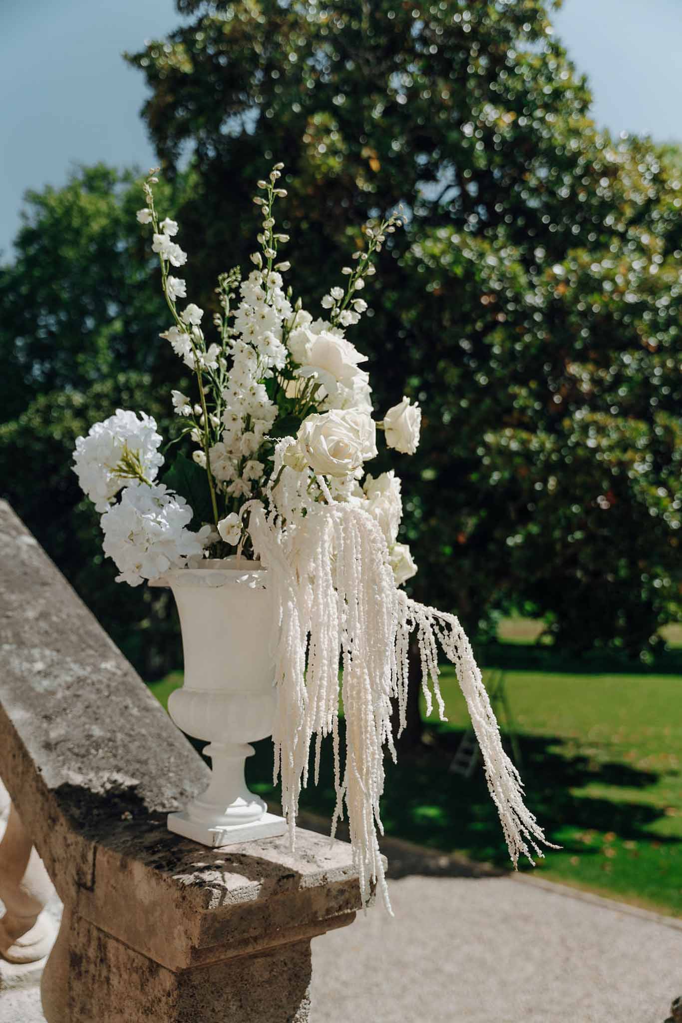 White pedestal urn with garden roses, delphinium, stock, and trailing amaranthus on stone balustrade in garden