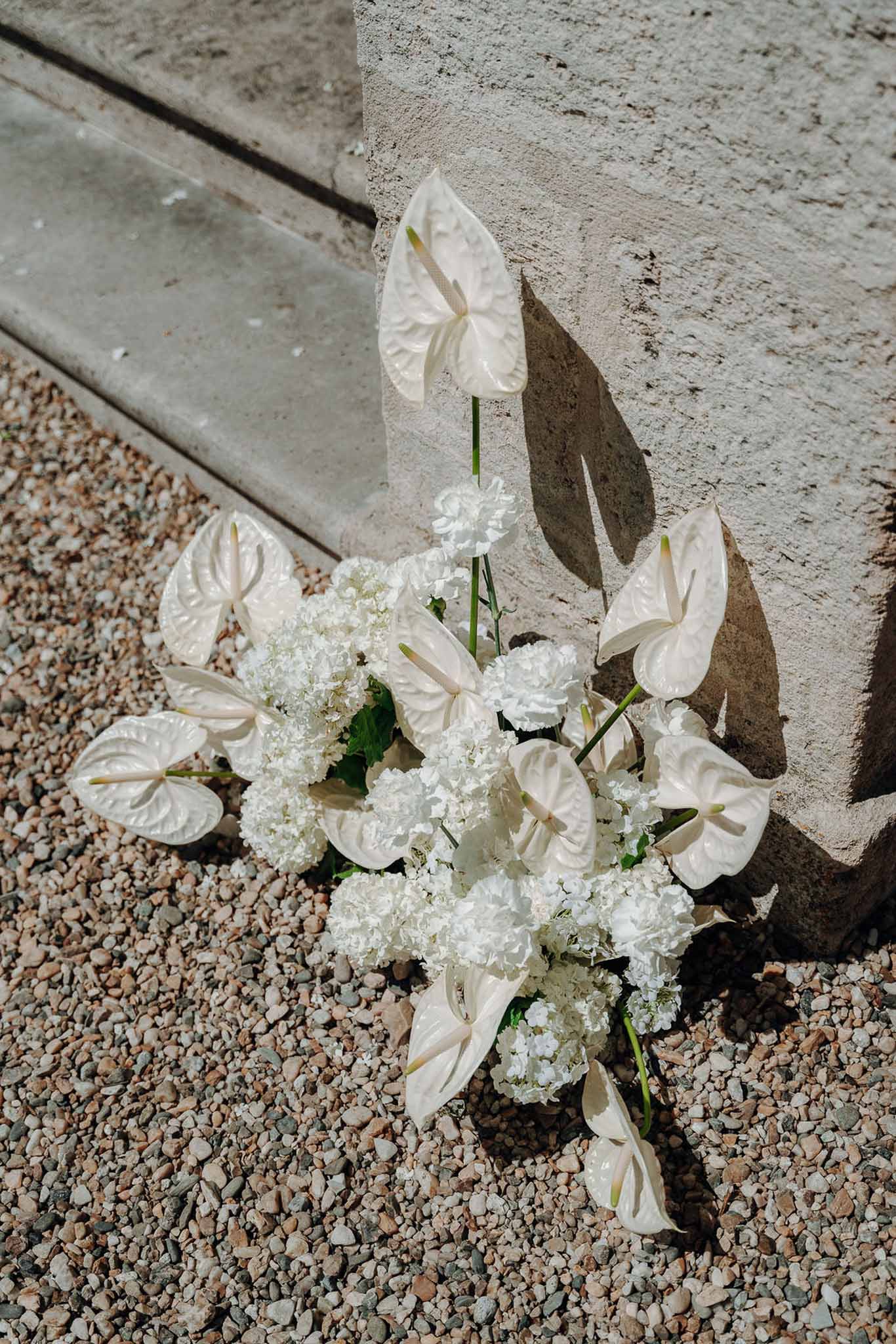 Close-up of white anthurium hydrangea and carnation floral arrangement on gravel beside stone architectural element