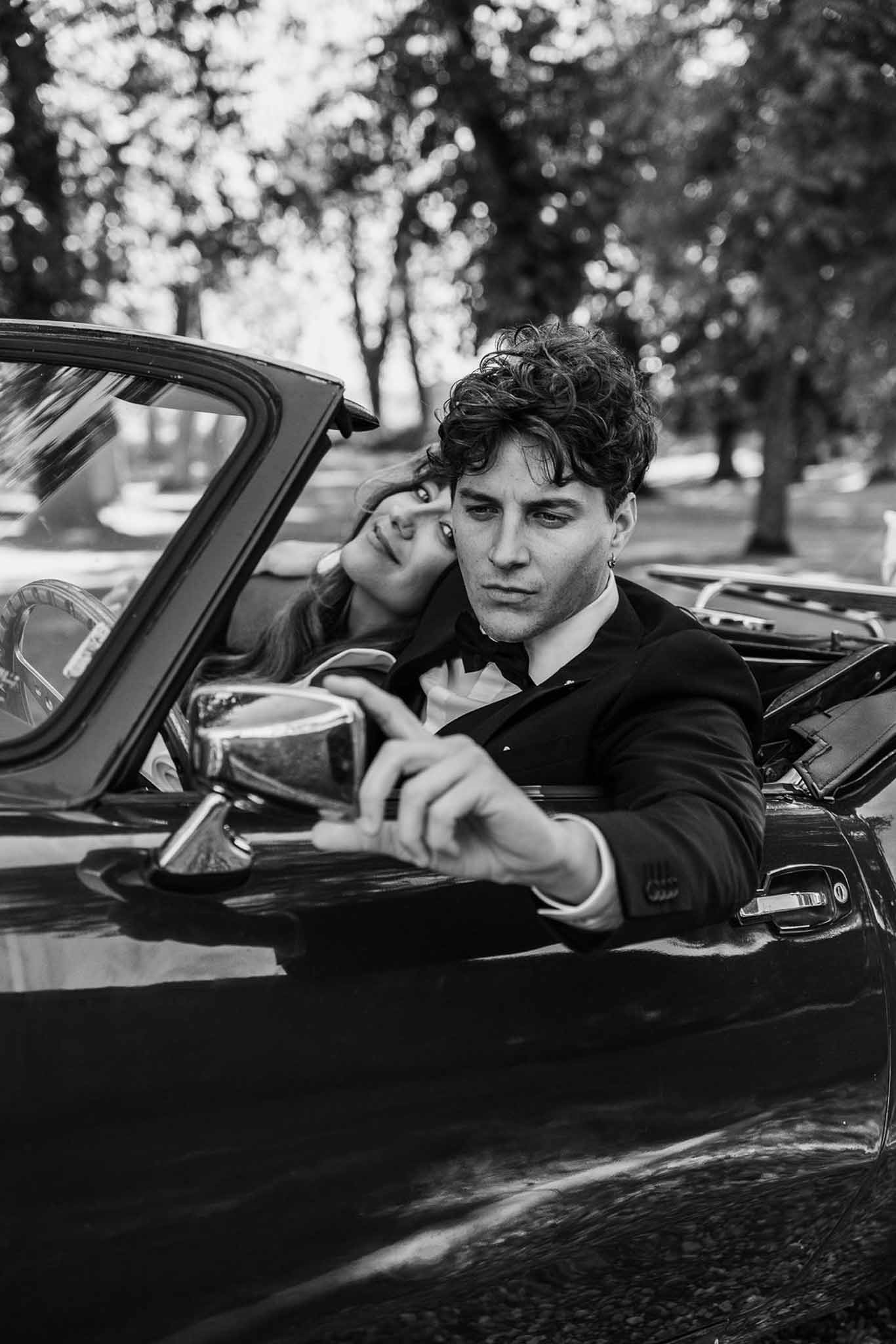 Black and white portrait of groom in vintage convertible with bride resting head on his shoulder