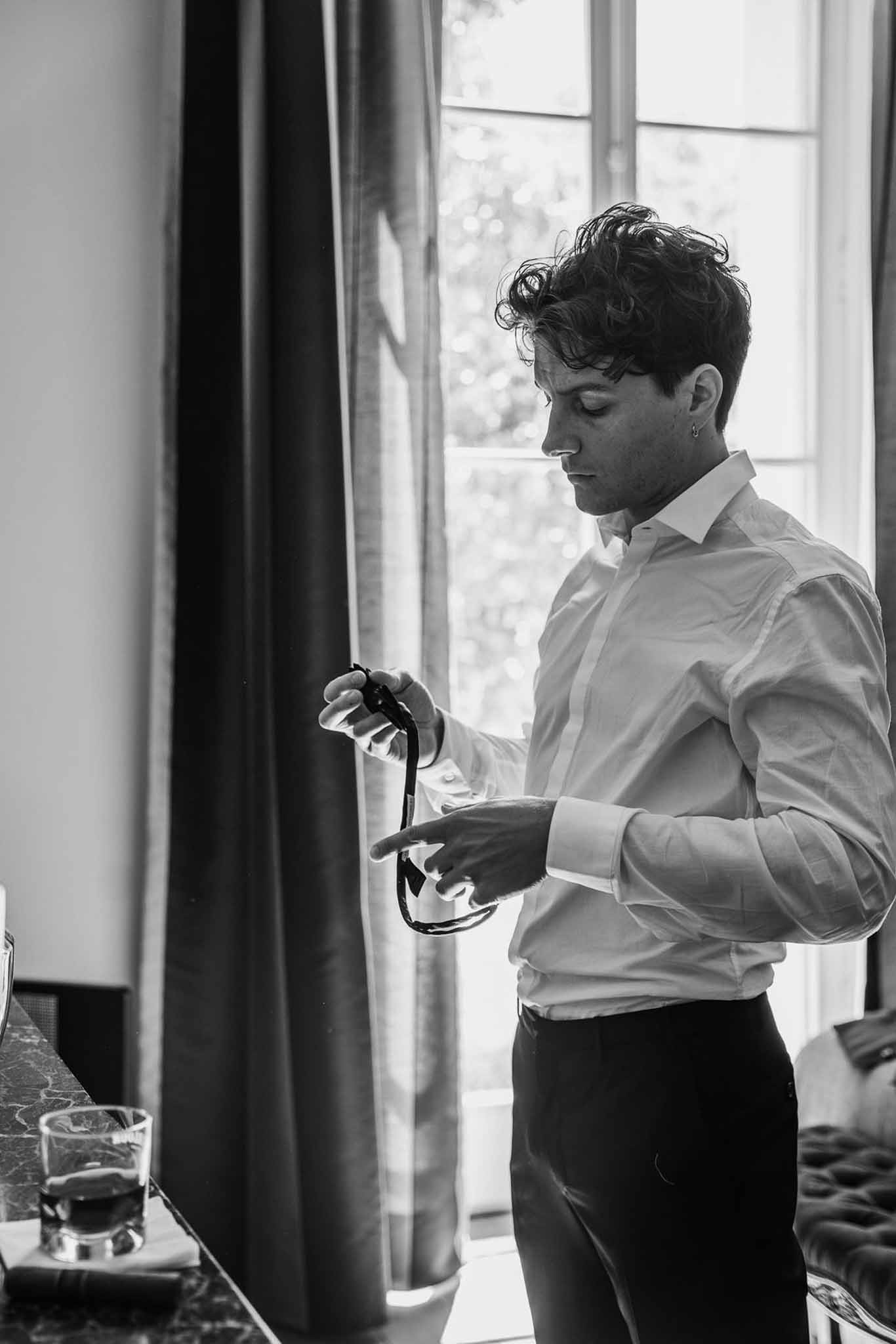 Black-and-white portrait of groom in white dress shirt examining bow tie by tall window with natural light
