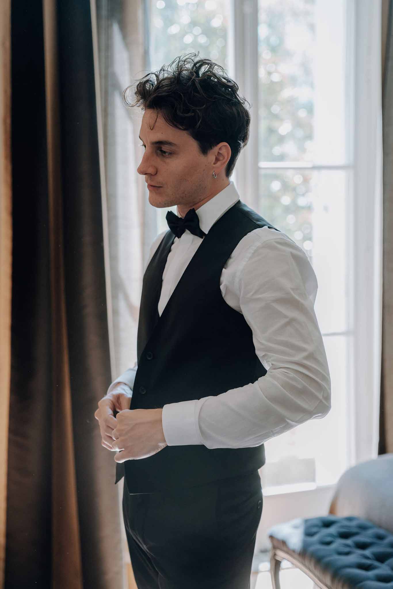 Groom buttoning black waistcoat with bow tie beside window in chateau room with warm drapes