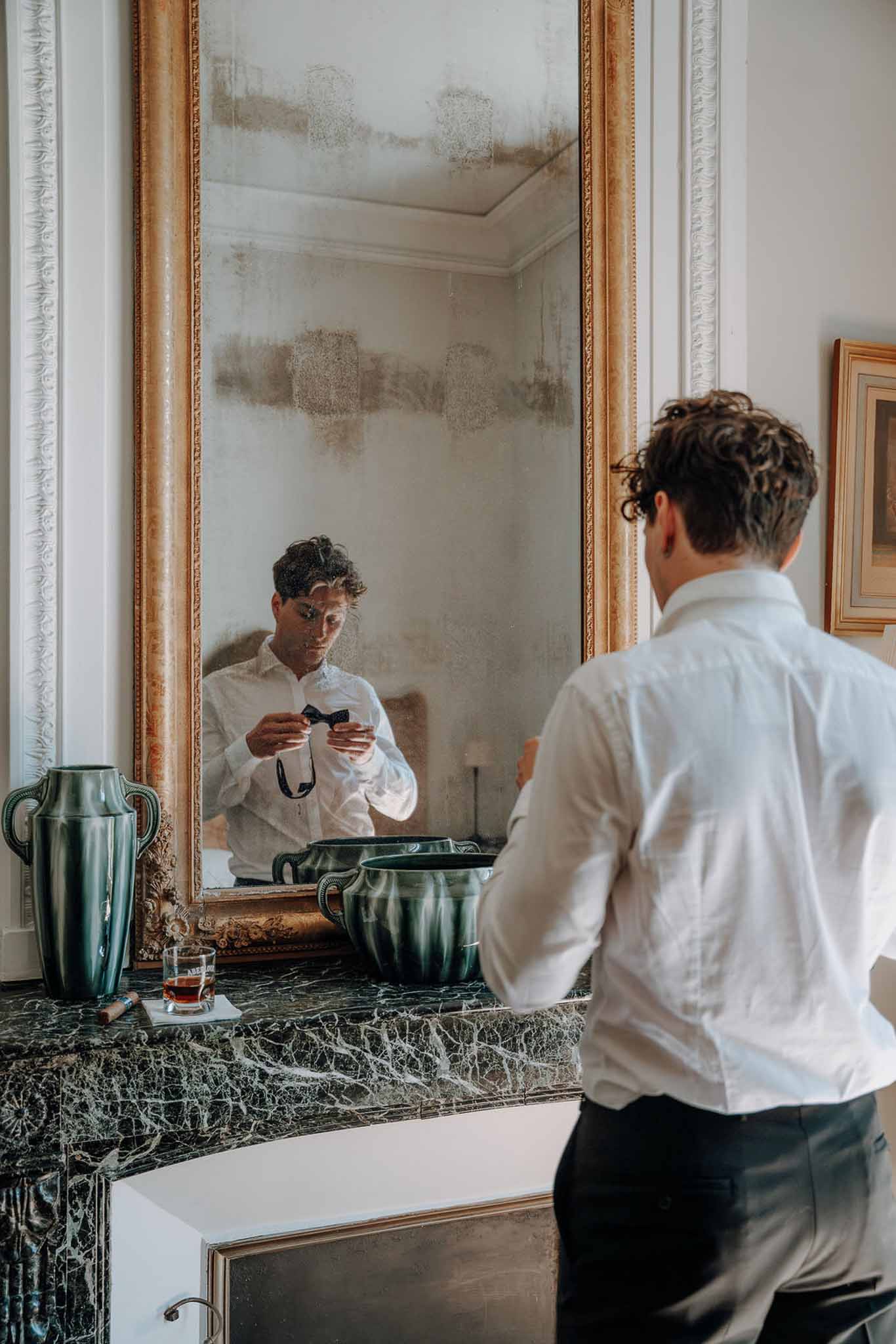 Groom fastening navy bow tie at dark green marble mantel reflected in gold-framed antique mirror