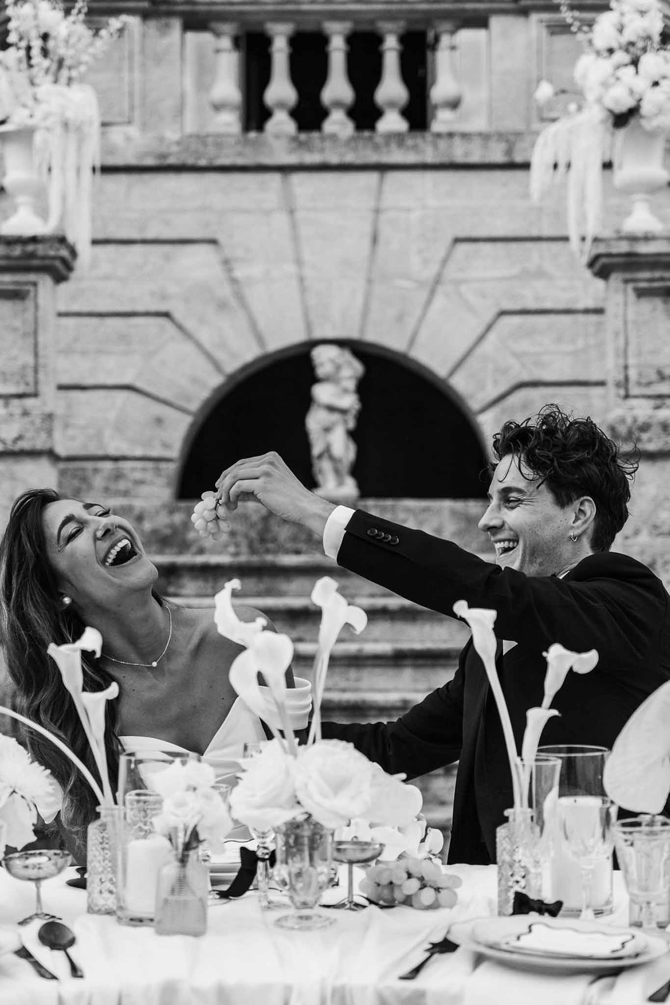 Black and white shot of bride and groom laughing at sweetheart table with calla lilies and chateau facade behind