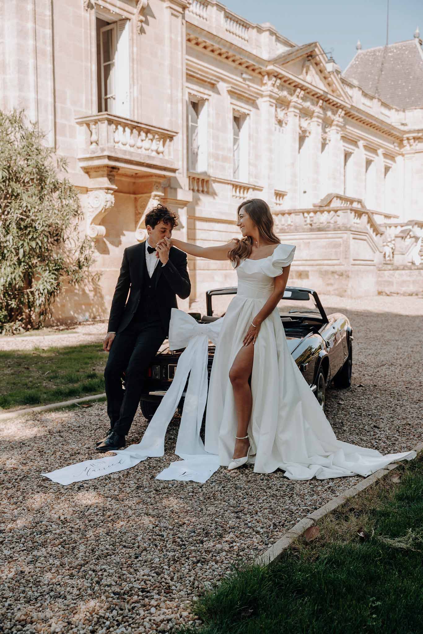 Groom kissing bride's hand beside vintage black convertible, bride in white gown with satin bow and calligraphy ribbon