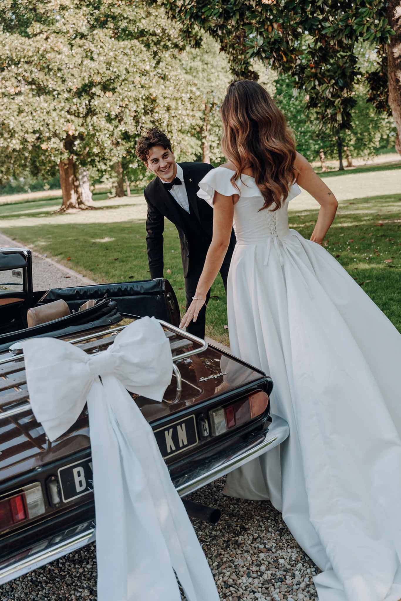 Bride and groom beside a vintage black convertible with white satin bow on gravel driveway in parkland grounds