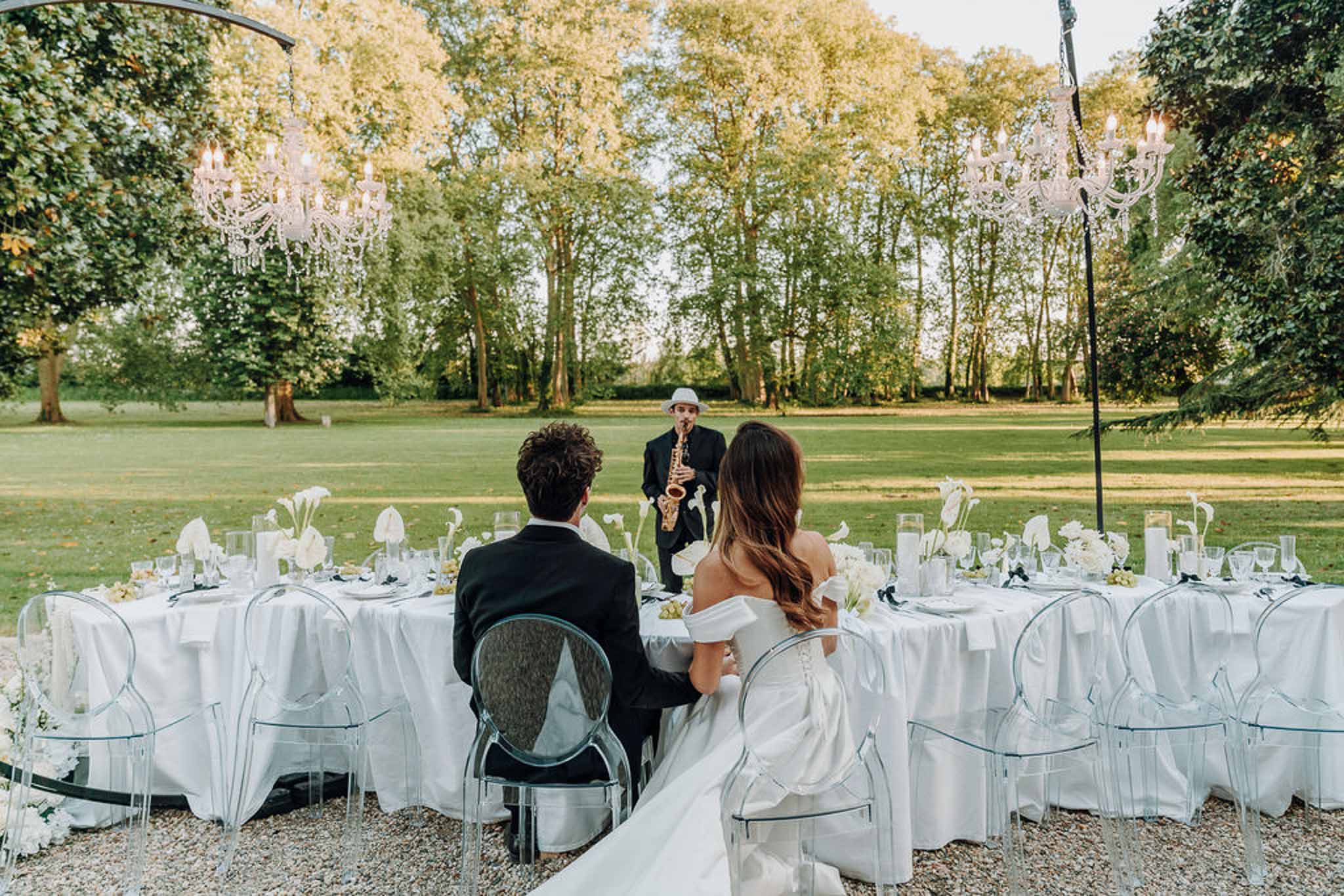 Bride and groom seated at outdoor reception table watching a saxophonist with crystal chandeliers overhead
