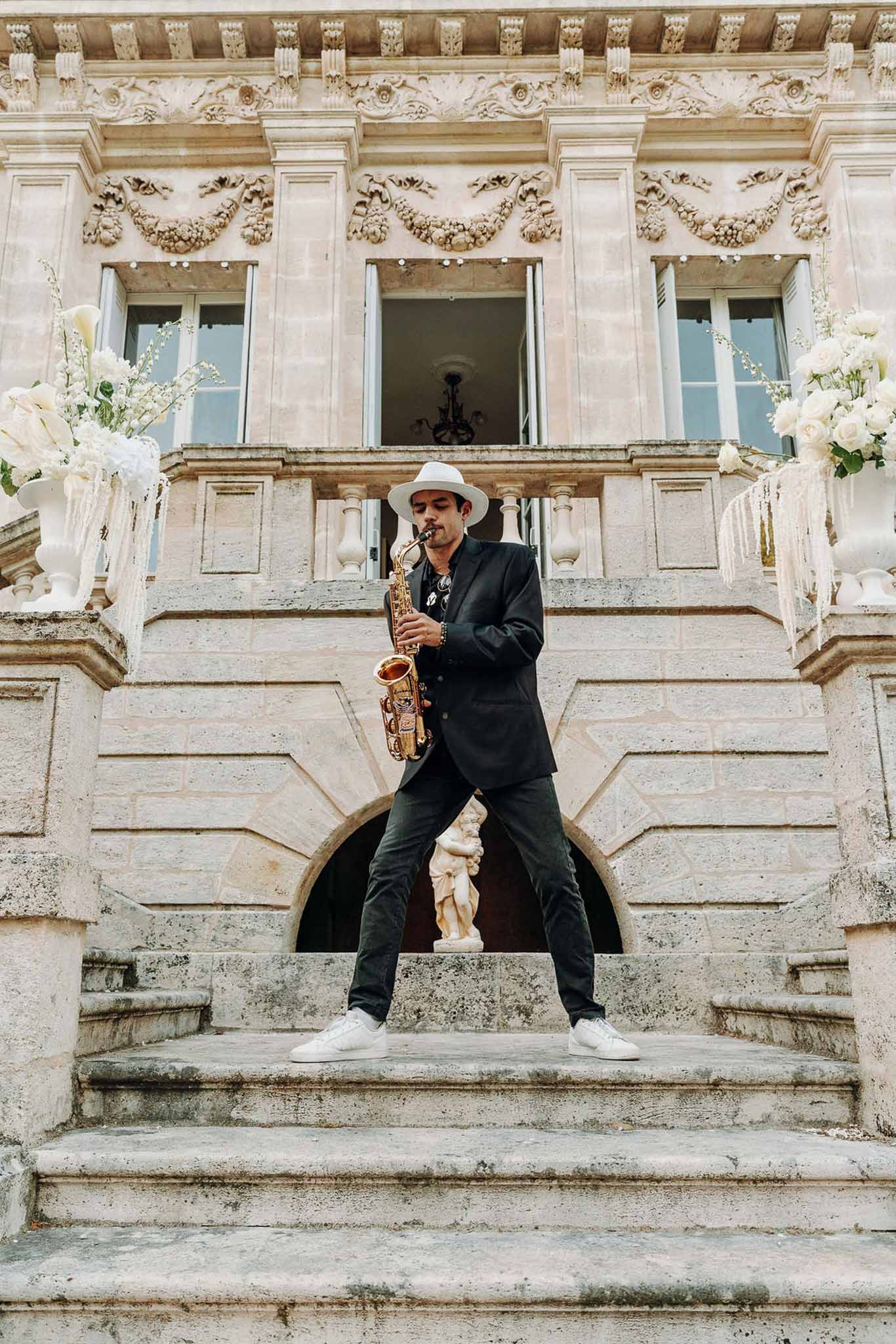 Saxophonist performing on chateau staircase flanked by white rose and calla lily arrangements on pedestals