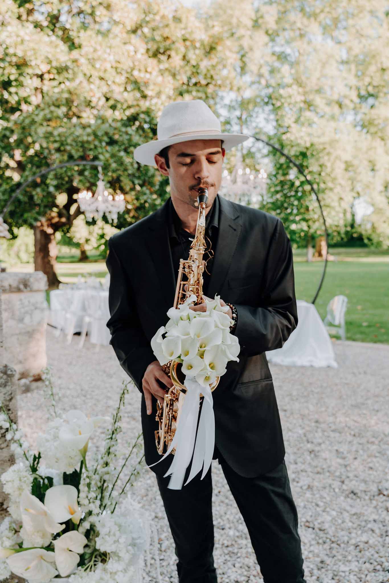 Saxophonist in black suit and white fedora with calla lily decorated gold saxophone before crystal chandelier arch