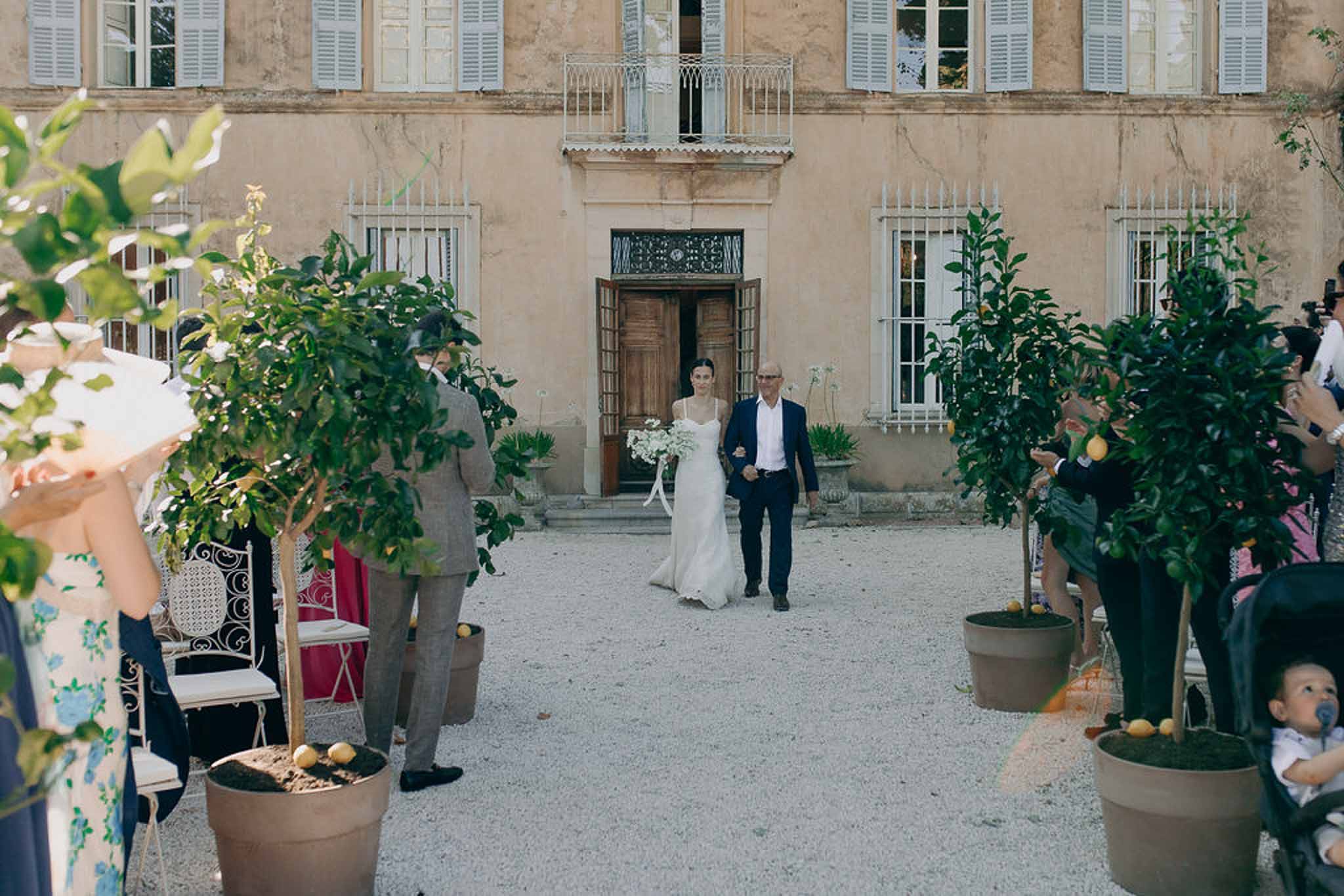 Bride walking down aisle with father past potted lemon trees in ochre chateau courtyard ceremony
