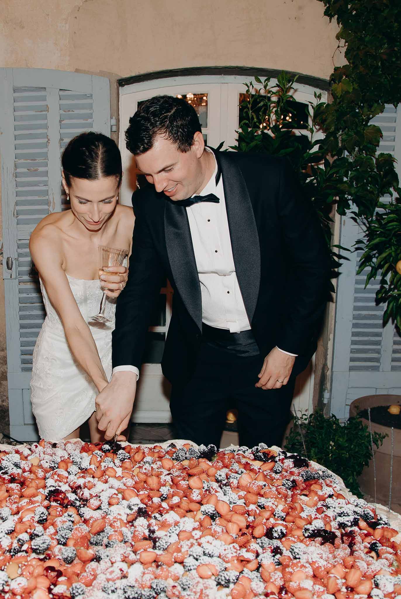 Couple cutting strawberry and berry topped tart at evening reception bride in white lace mini dress groom in tuxedo