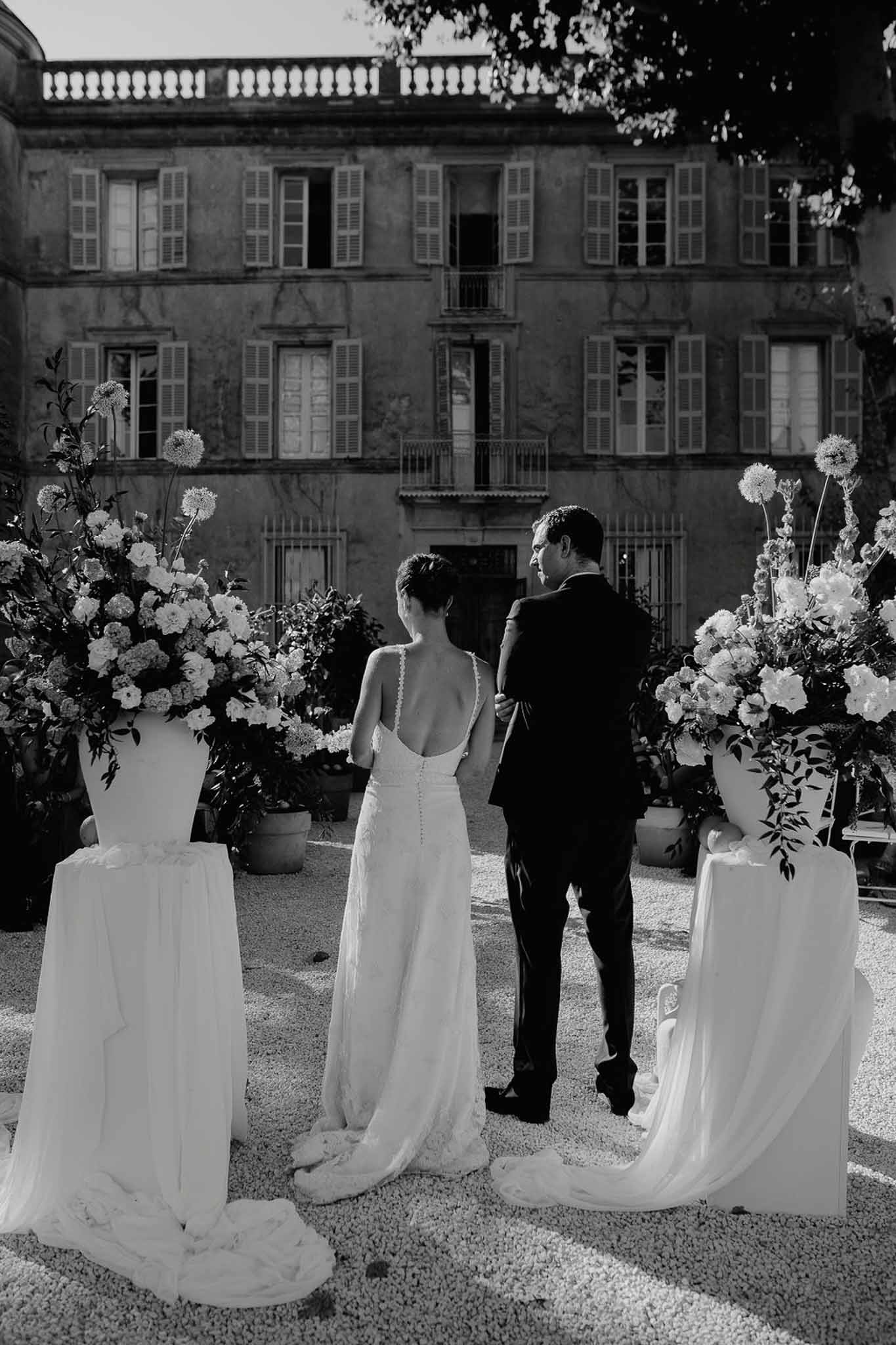 Bride and groom from behind between white floral plinths facing chateau facade during ceremony in B&W