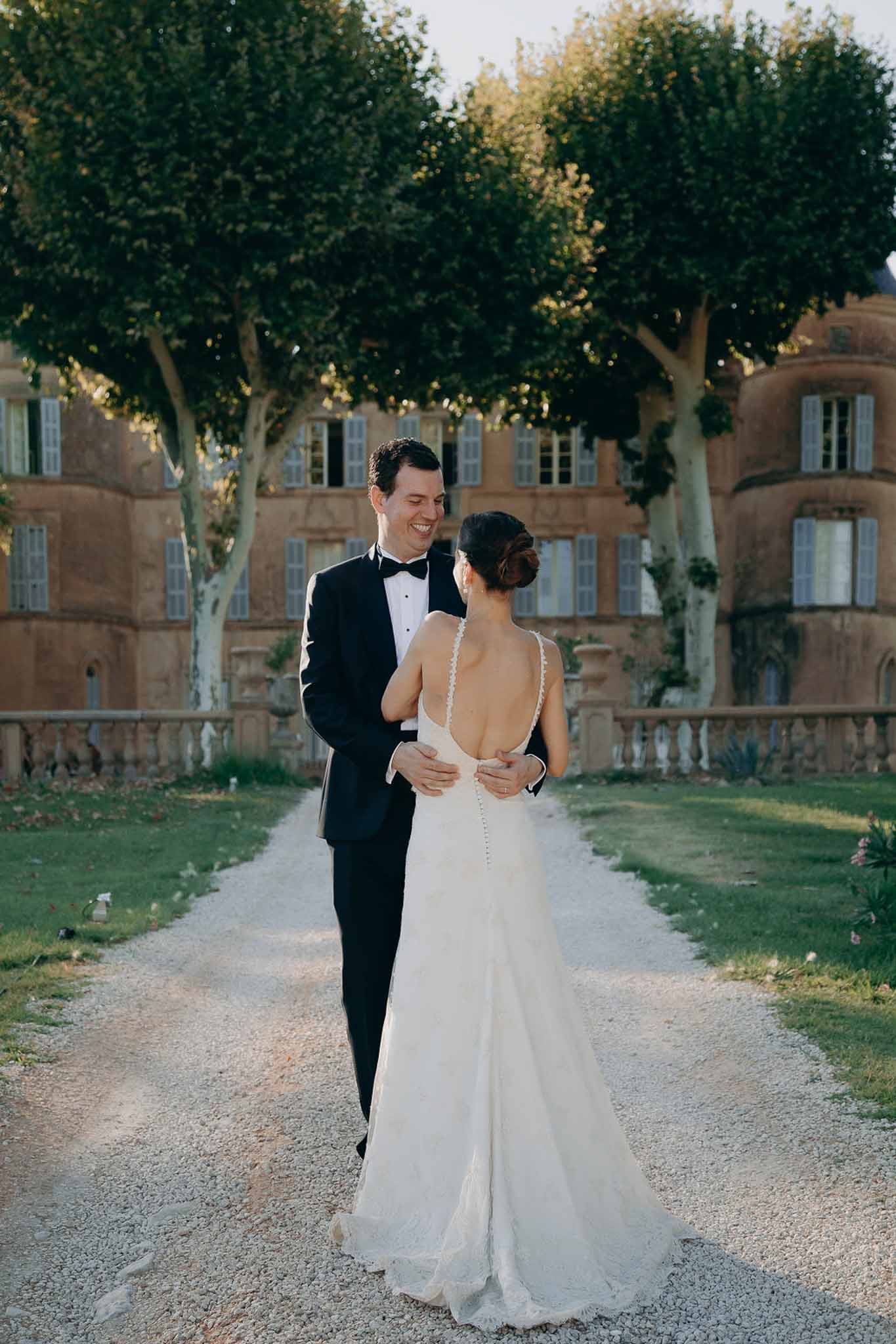 Groom smiling at camera while holding bride showing open-back lace gown in front of ochre chateau