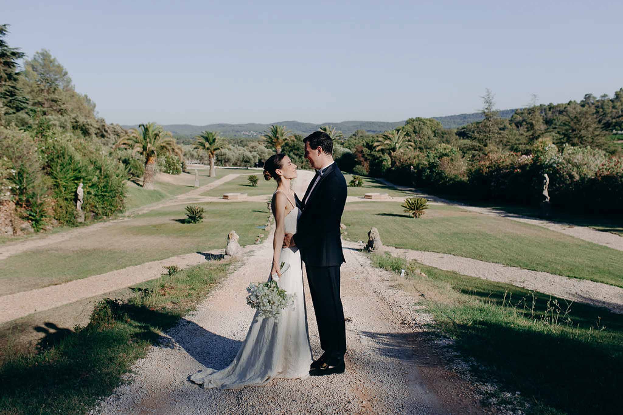 Couple facing each other in formal garden with statues and palm trees, bride with blue and white bouquet