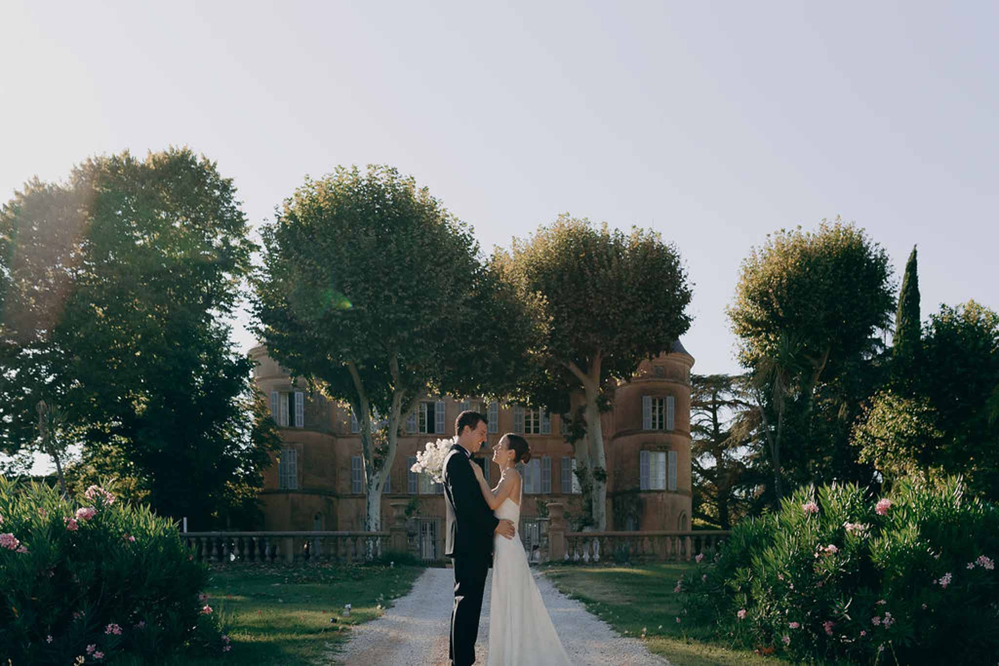 Bride and groom standing forehead to forehead on chateau driveway lined with pink flowering shrubs