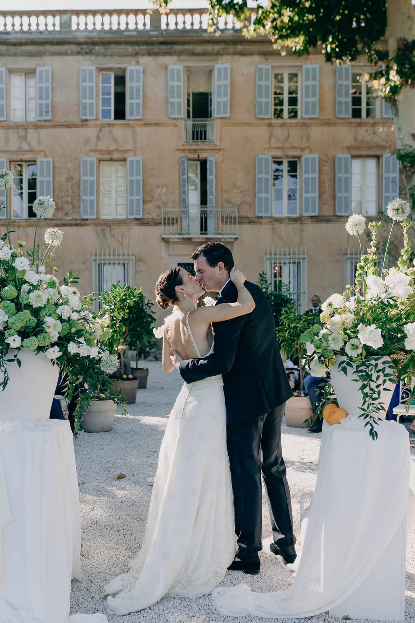 Bride and groom share first kiss at outdoor ceremony in Provencal chateau courtyard flanked by white rose urns