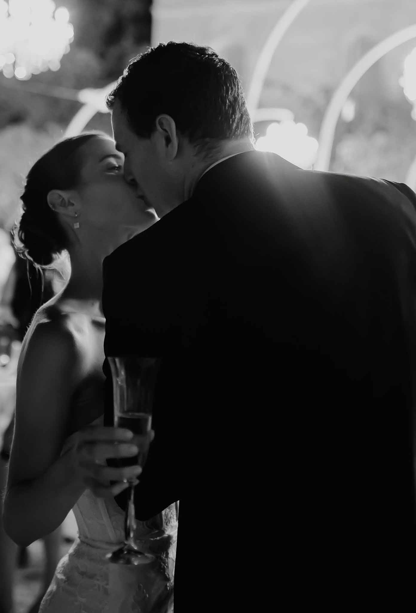 Backlit close-up of couple kissing with champagne flute in hand at indoor reception in B&W